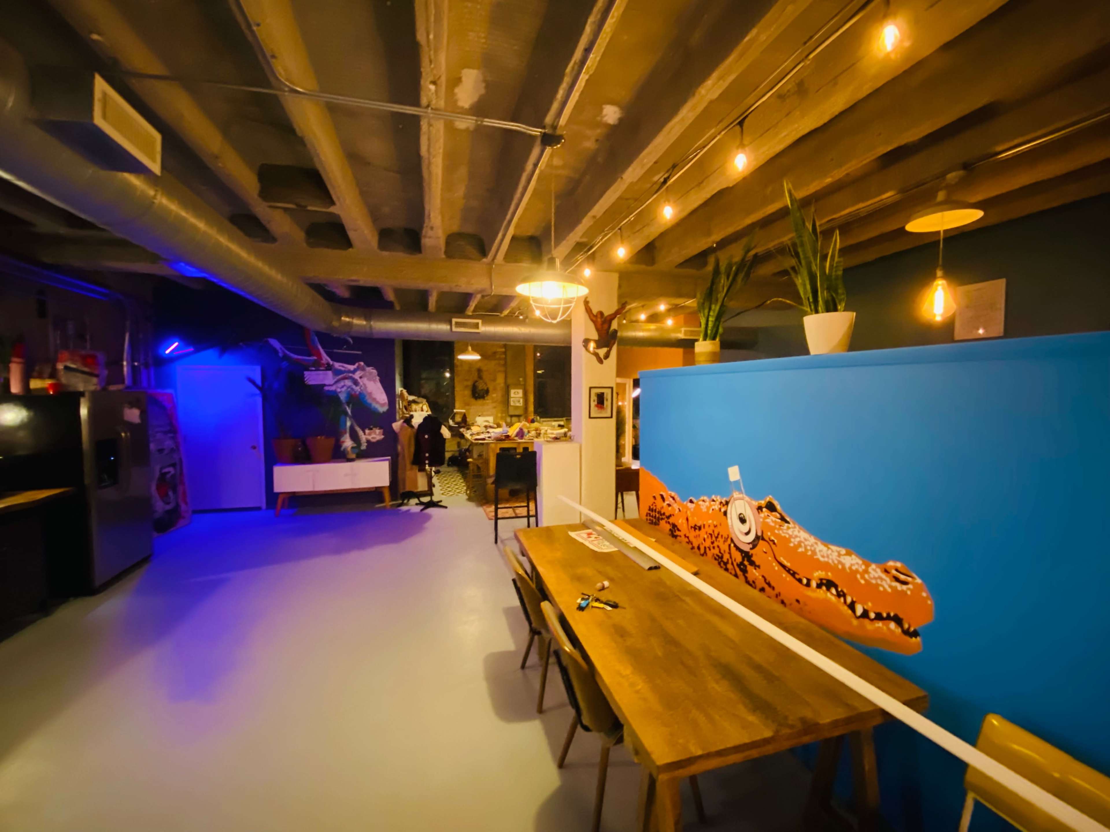 The image shows a modern interior space with a wooden table, colorful wall art featuring a crocodile, and a kitchen area to the side.