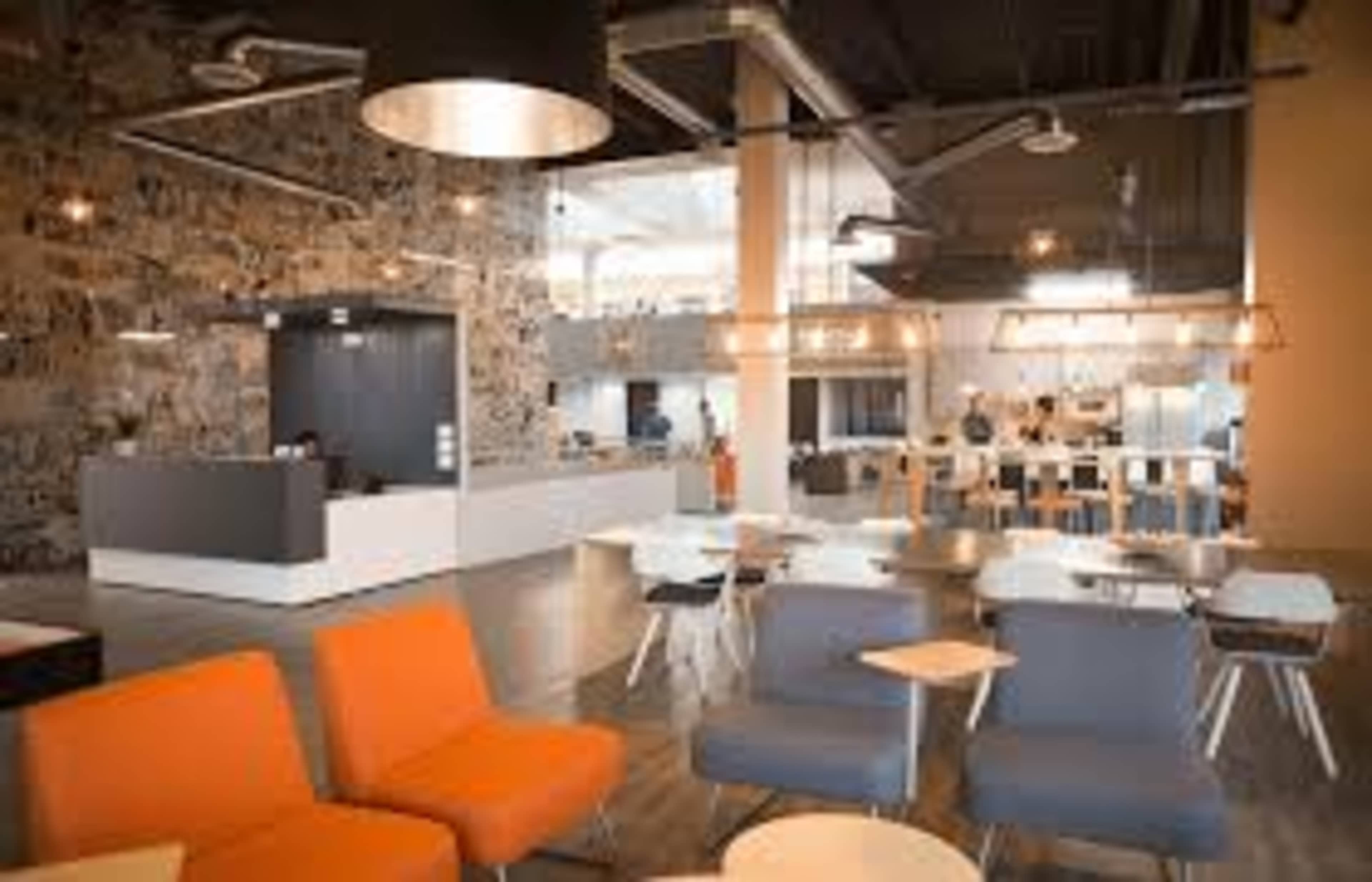 A modern office interior featuring a reception area with a stone wall, seating arrangements with orange chairs, and a kitchen space in the background.