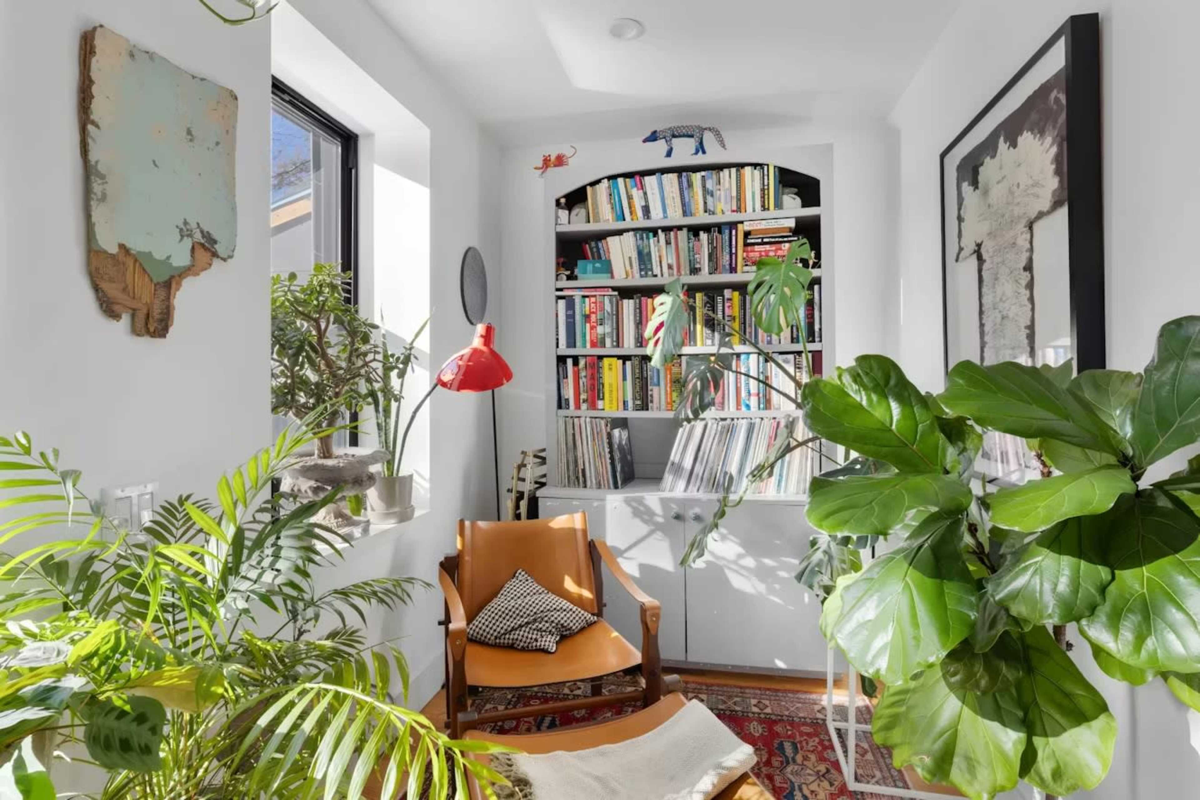 A cozy reading nook features a chair, a vibrant lamp, and a bookshelf filled with books, surrounded by various indoor plants and natural light.