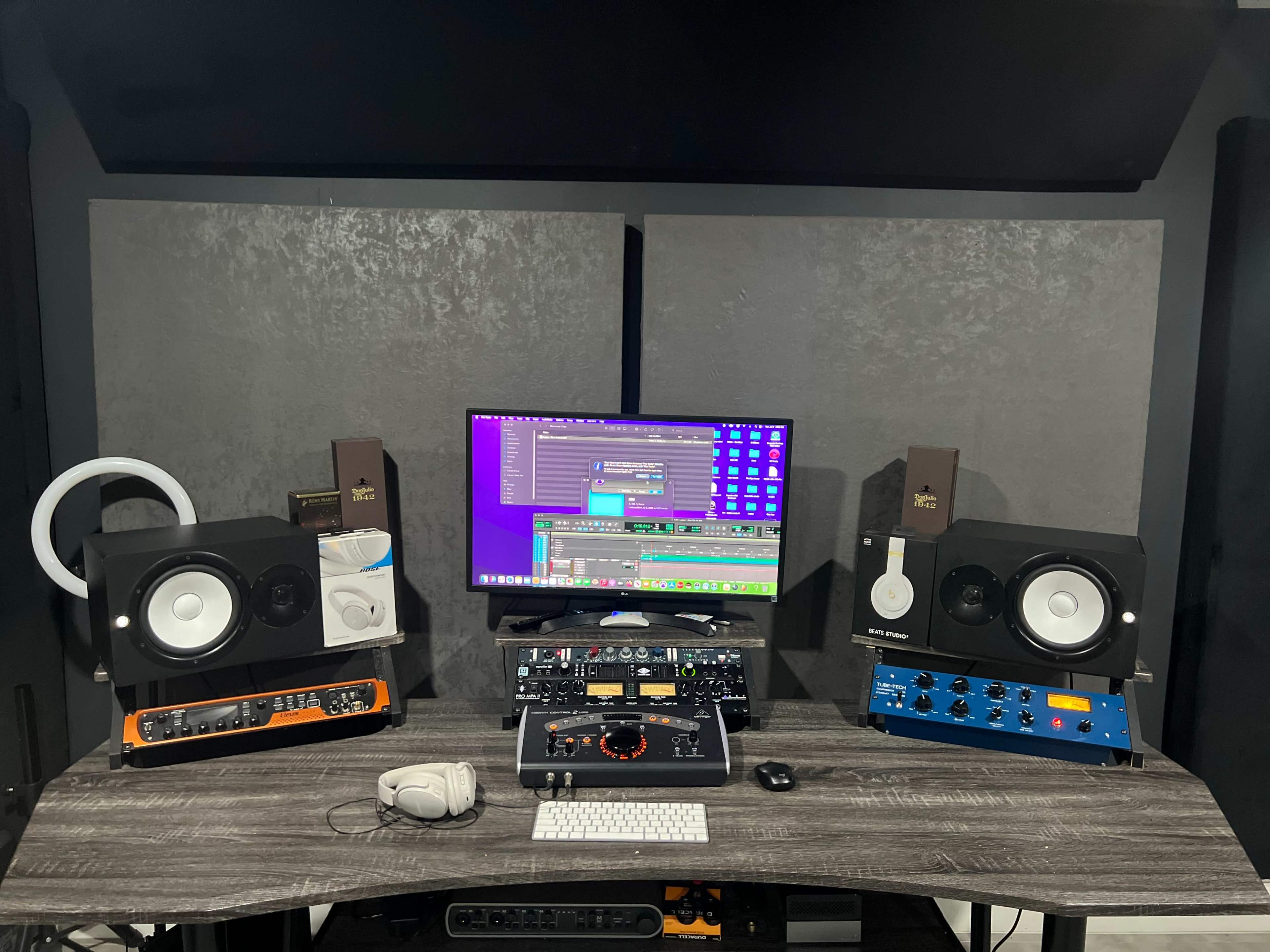 A home studio setup features a computer monitor, audio speakers, and various audio processing equipment on a desk against a soundproof wall.