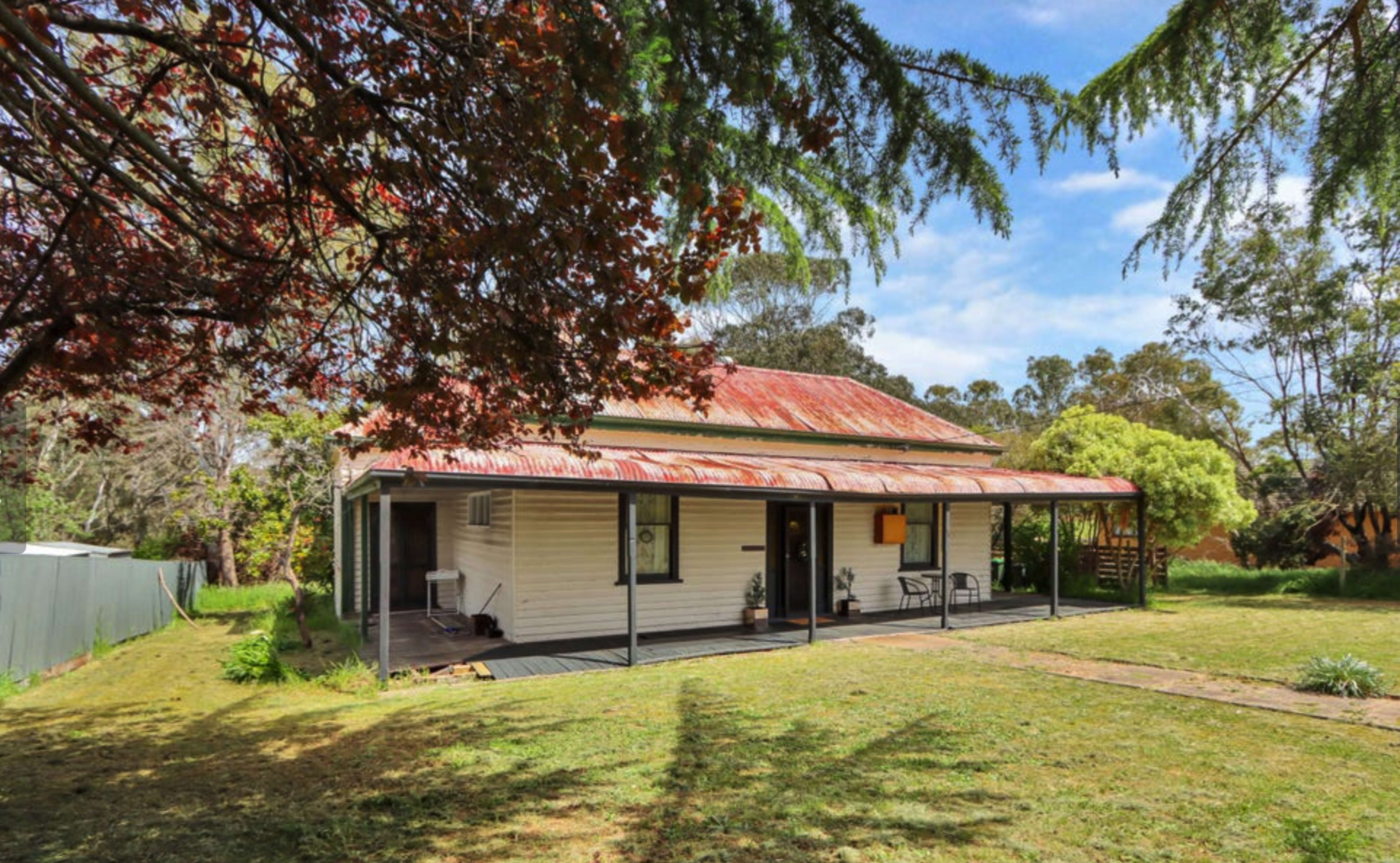 A single-story house with a rusted metal roof is surrounded by a grassy yard and trees.