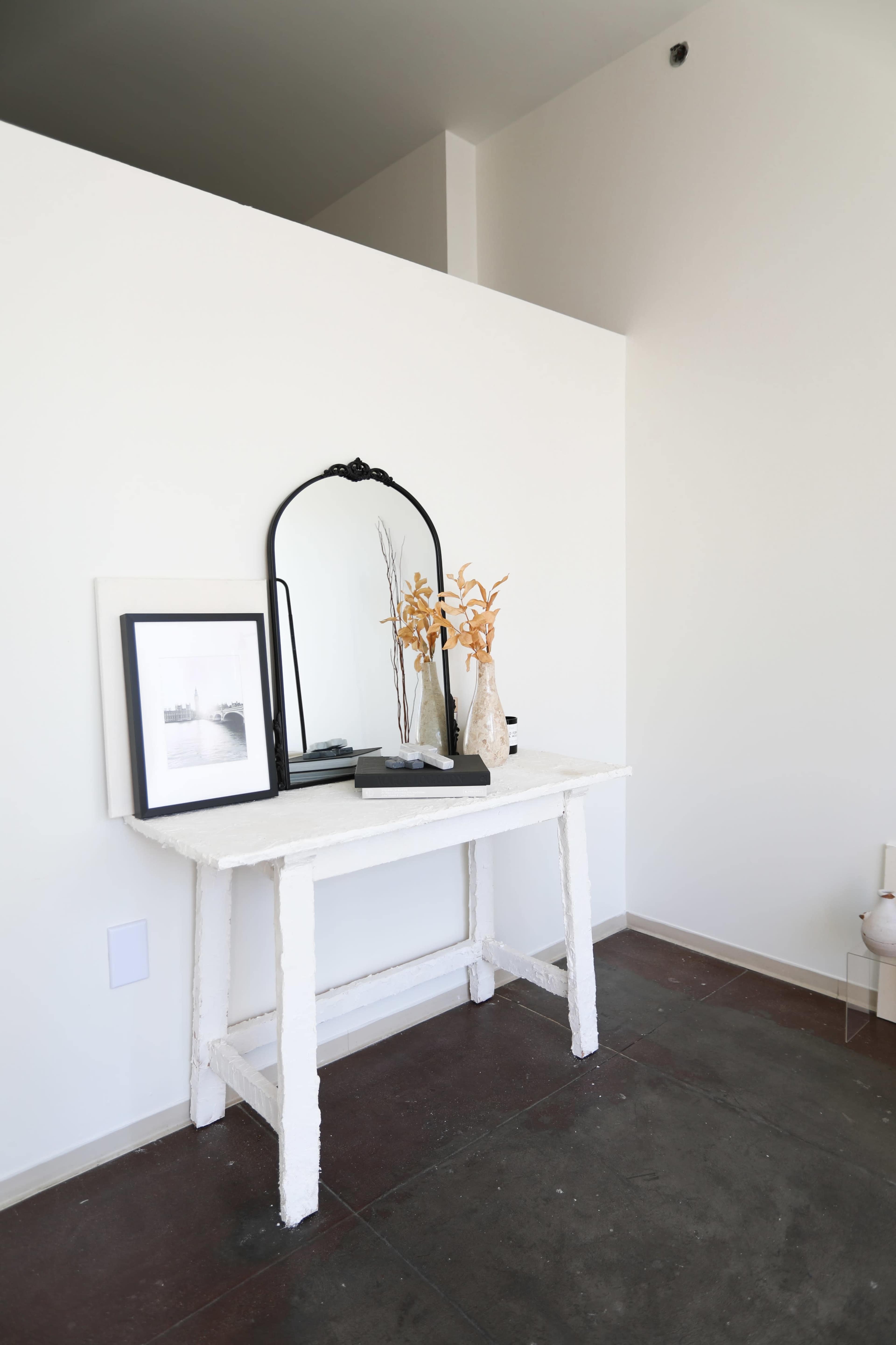 A simple white table with a mirror and decorative items is positioned against a light-colored wall.