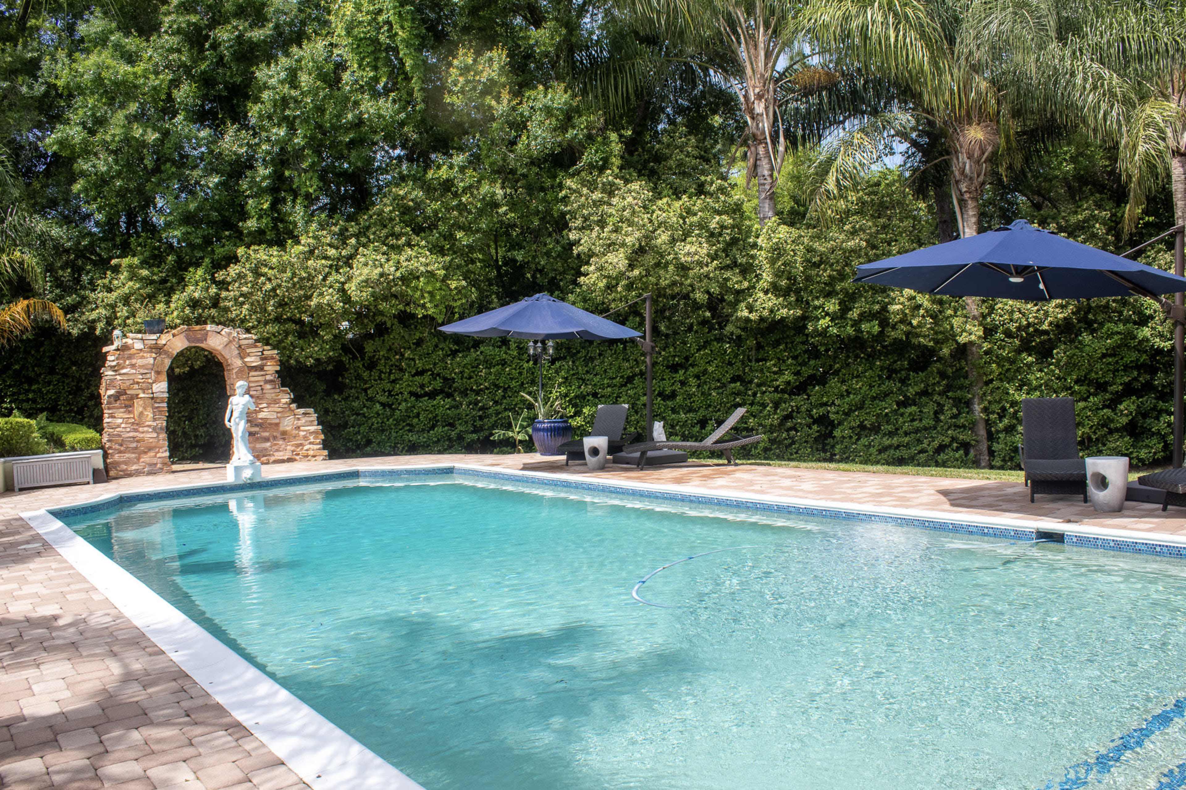 A landscaped backyard features a swimming pool with a stone archway and lounge chairs under blue umbrellas.