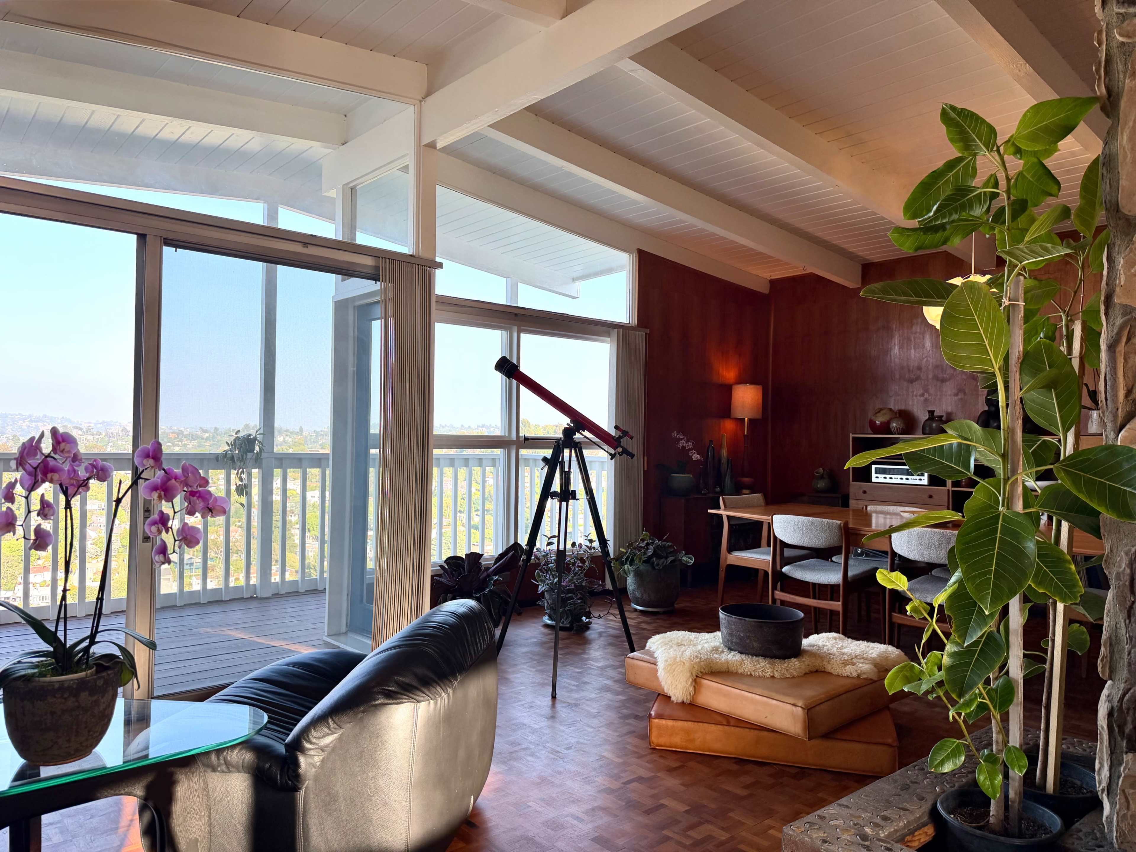 The image shows a spacious living area with large windows that provide a view of the outdoors, featuring a telescope, plants, and modern furniture.