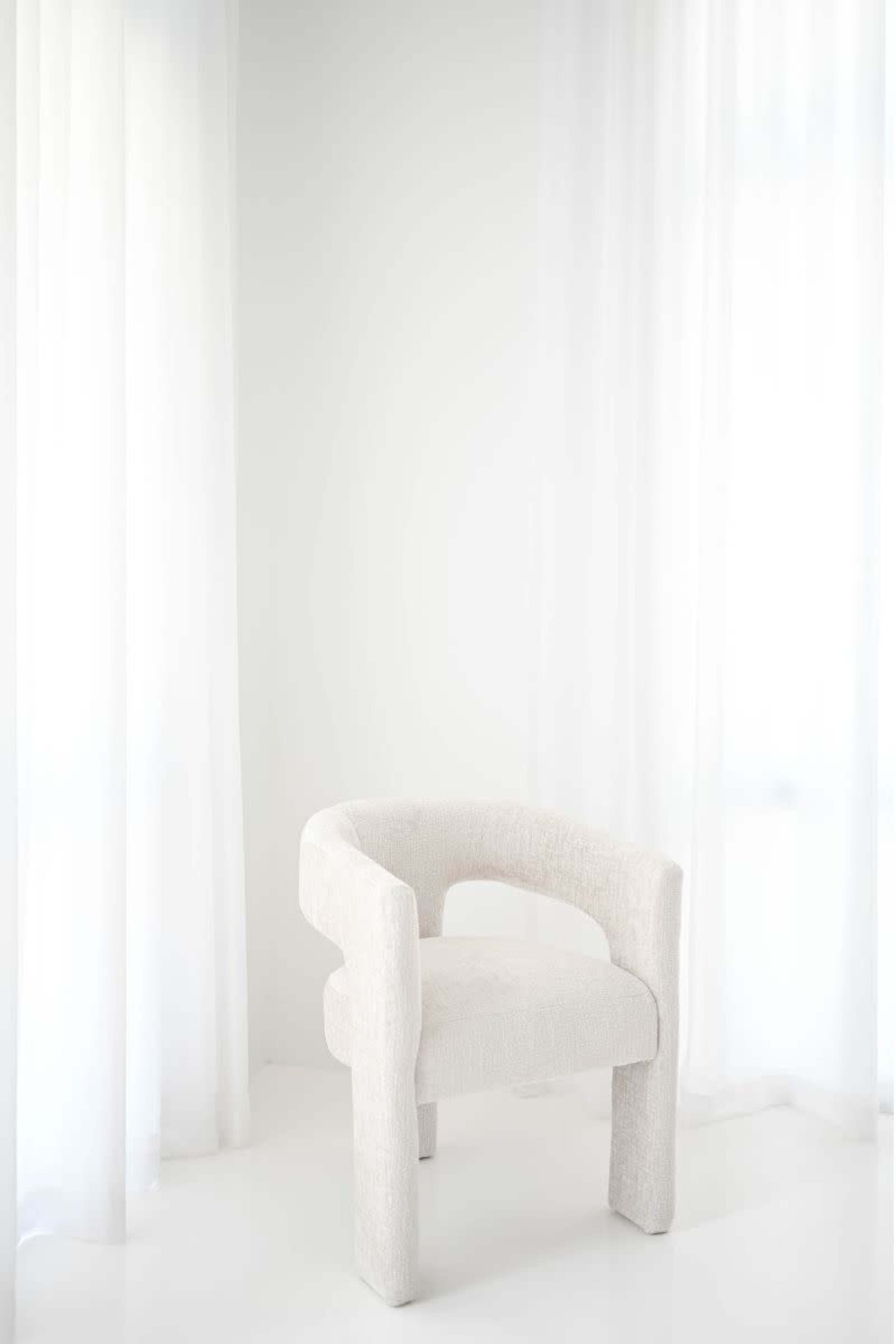 A light-colored, textured chair is positioned near sheer white curtains in a minimalist room with bright lighting.