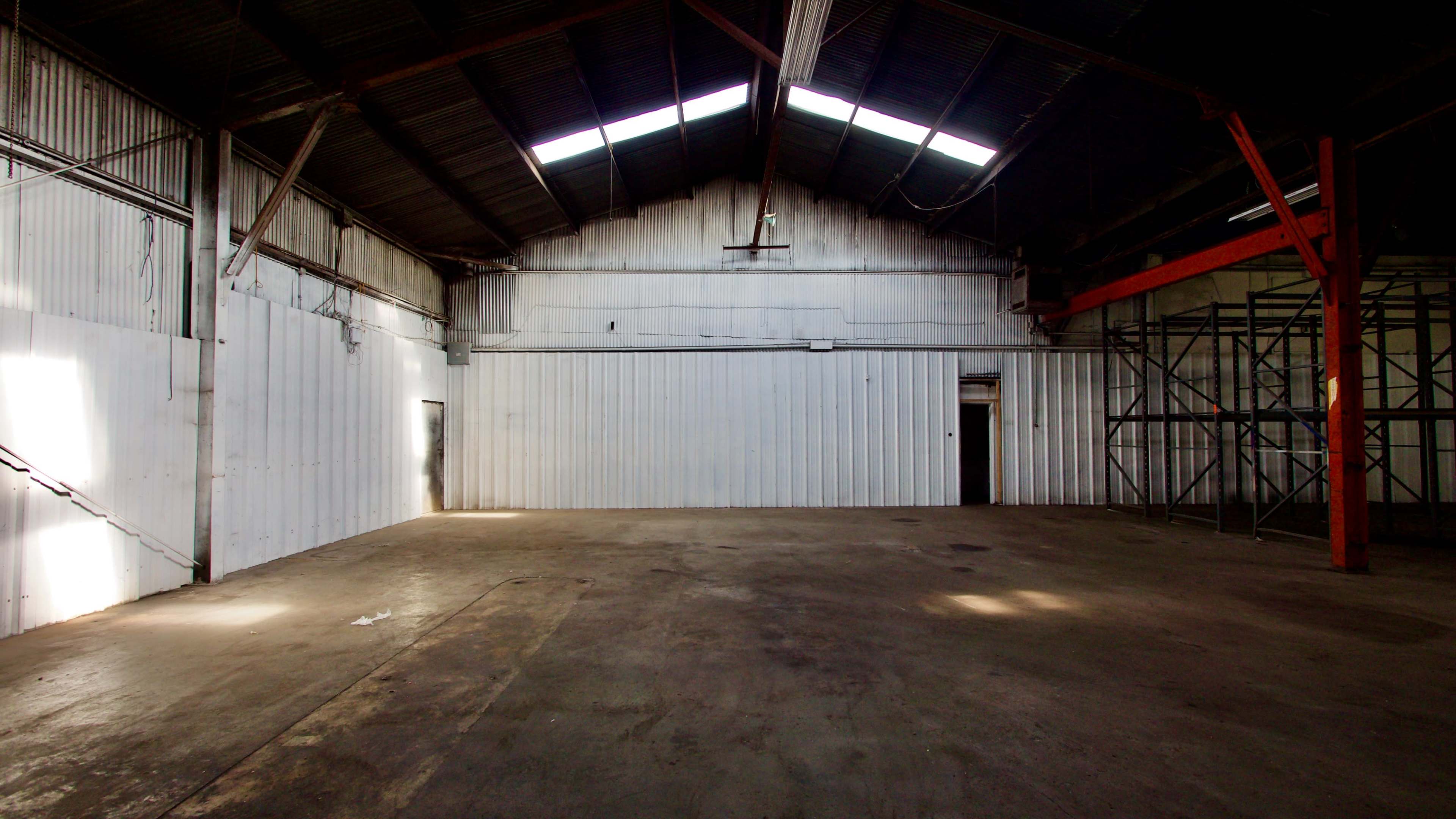 An empty warehouse with metal walls, a concrete floor, and a high ceiling featuring skylights.