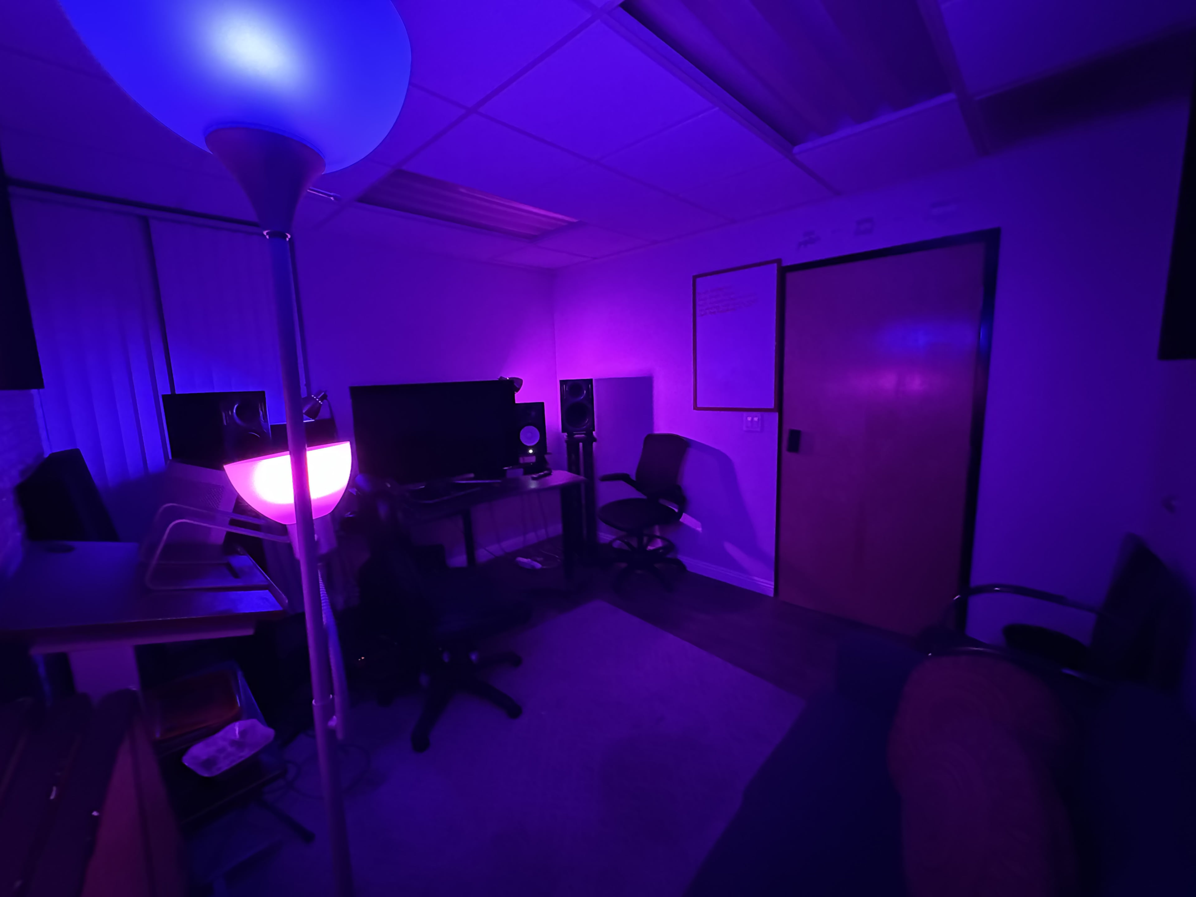 A dimly lit room with purple lighting features a desk with computer equipment, two chairs, and a whiteboard on the wall.