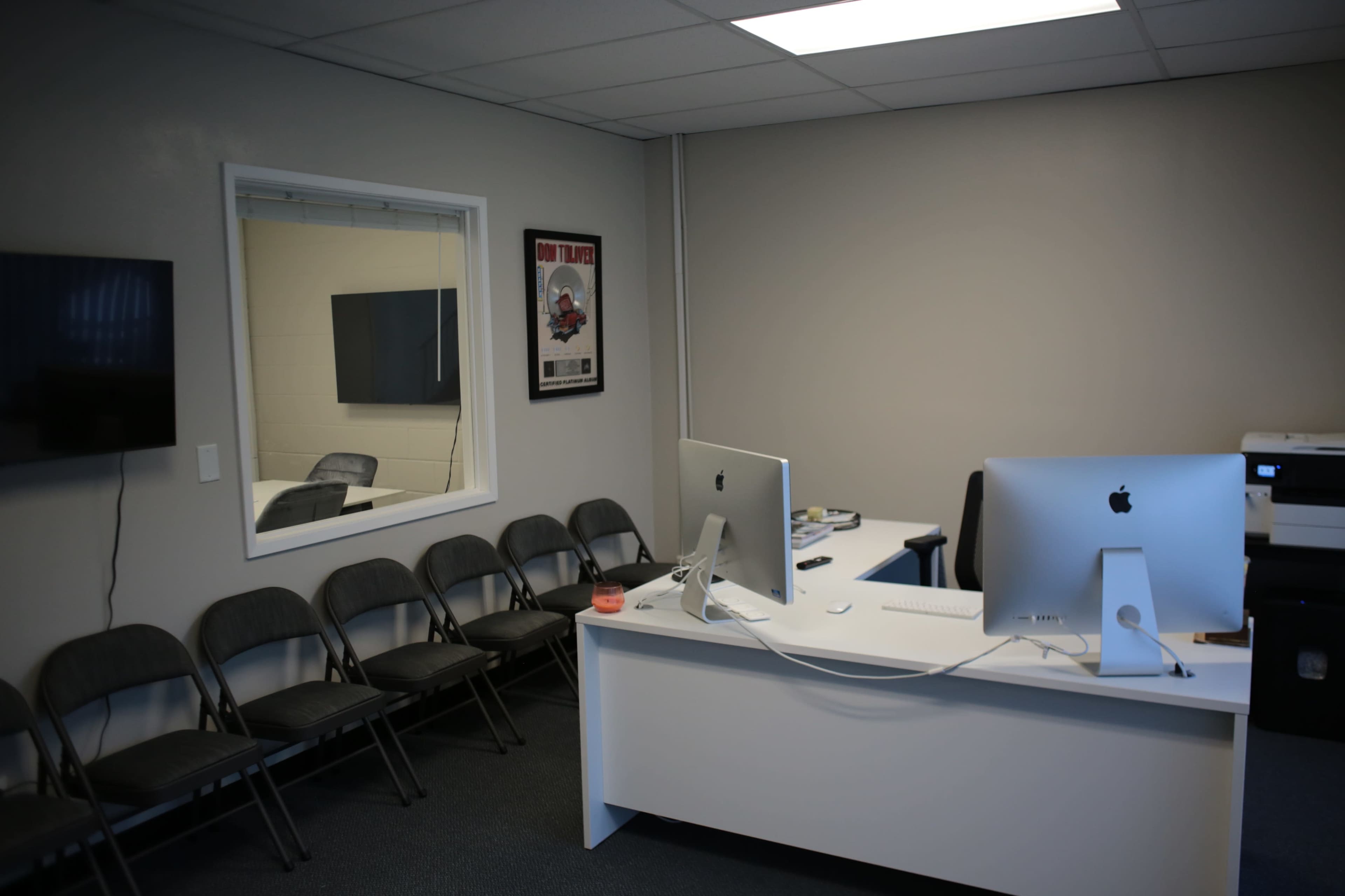 A minimalistic office with two Apple computers on a desk, a printer, and several folding chairs lined against the wall.