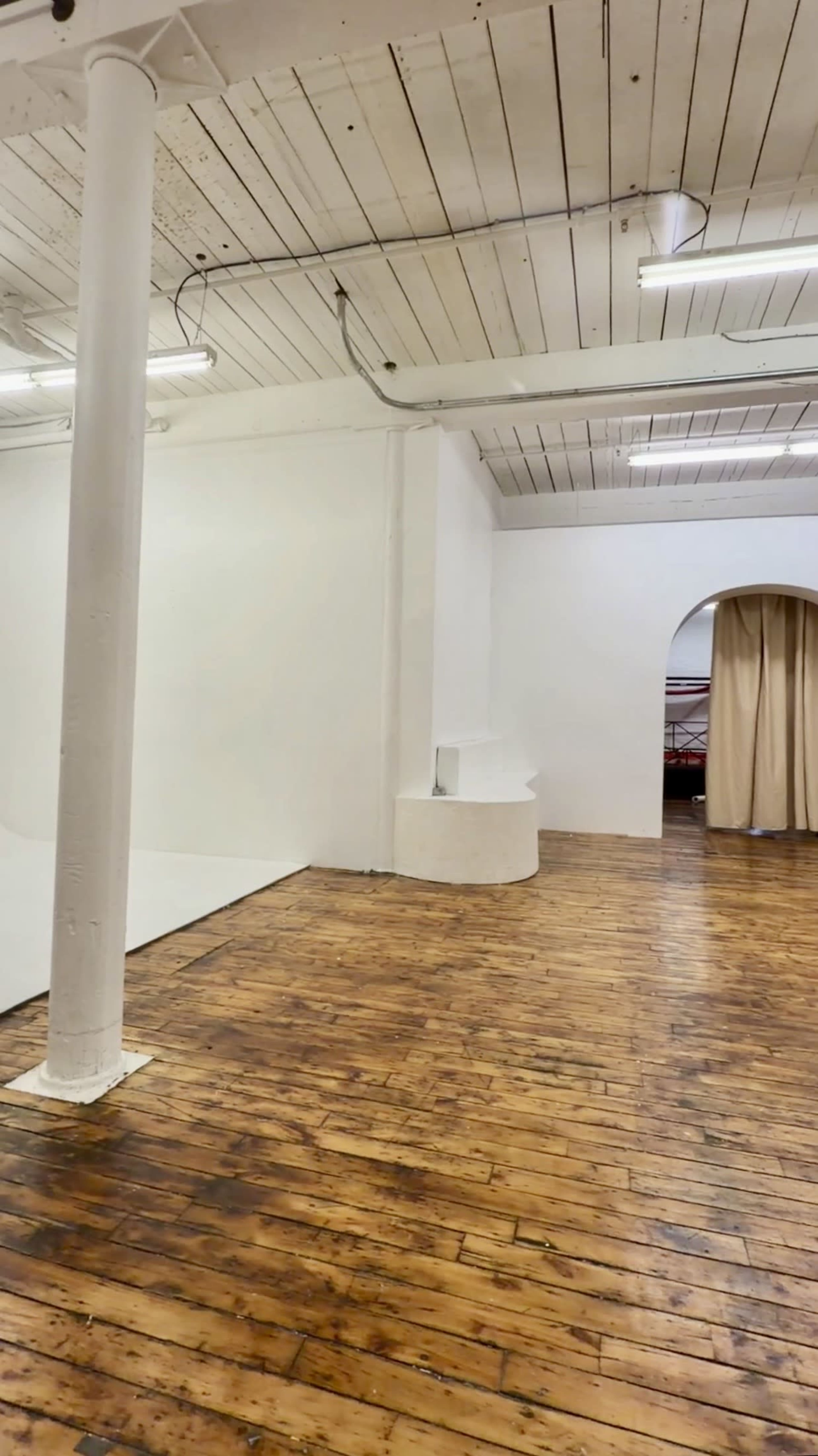 The image shows an empty indoor space with wooden floors, white walls, and a large archway leading to a curtained area.