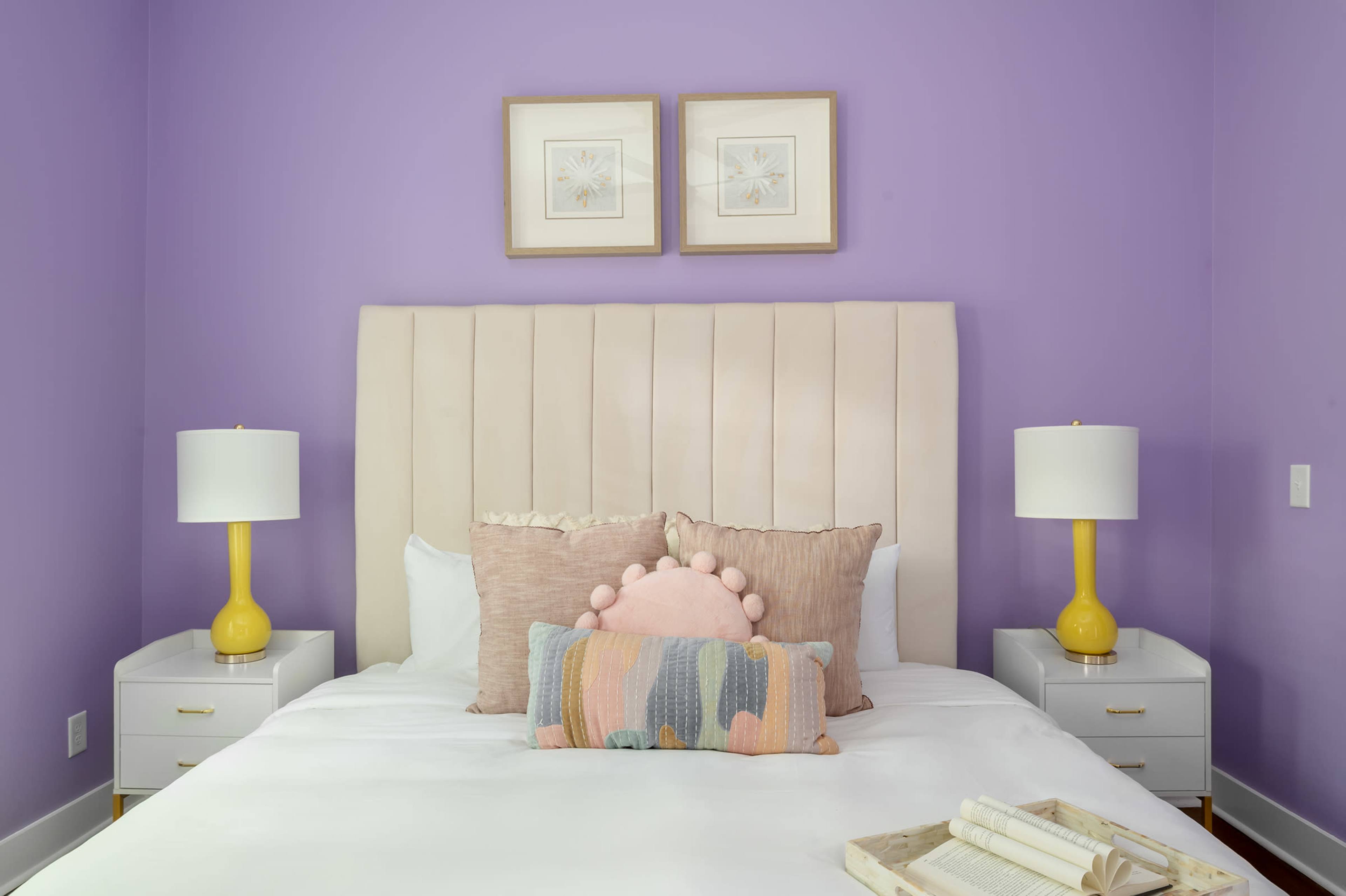 The image features a neatly made bed with a cream-colored headboard, colorful decorative pillows, and two yellow bedside lamps, all set against a lavender wall.