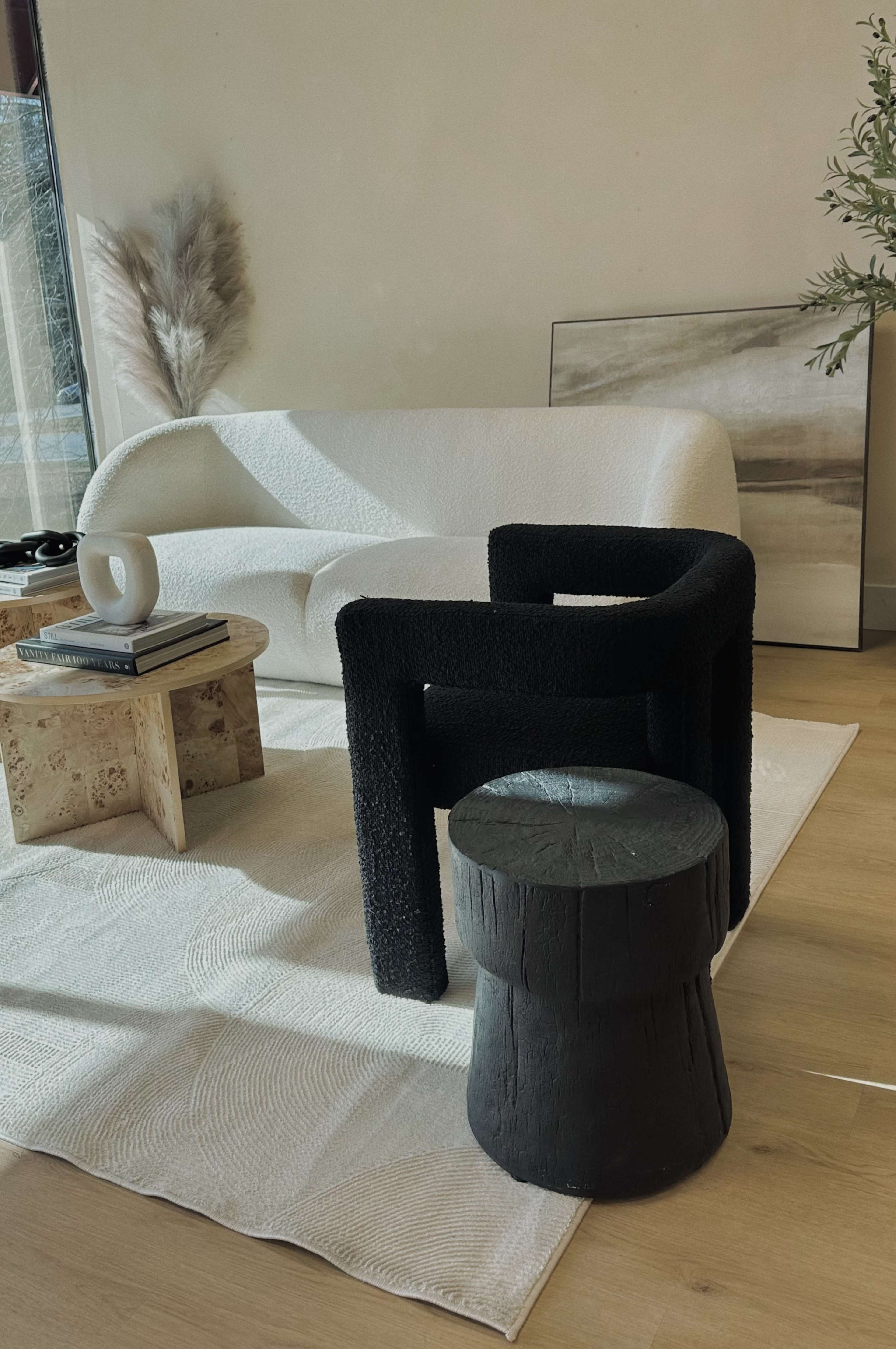 A textured black chair and a round black stool are positioned in a minimalist living space featuring a white sofa and a natural wood coffee table.