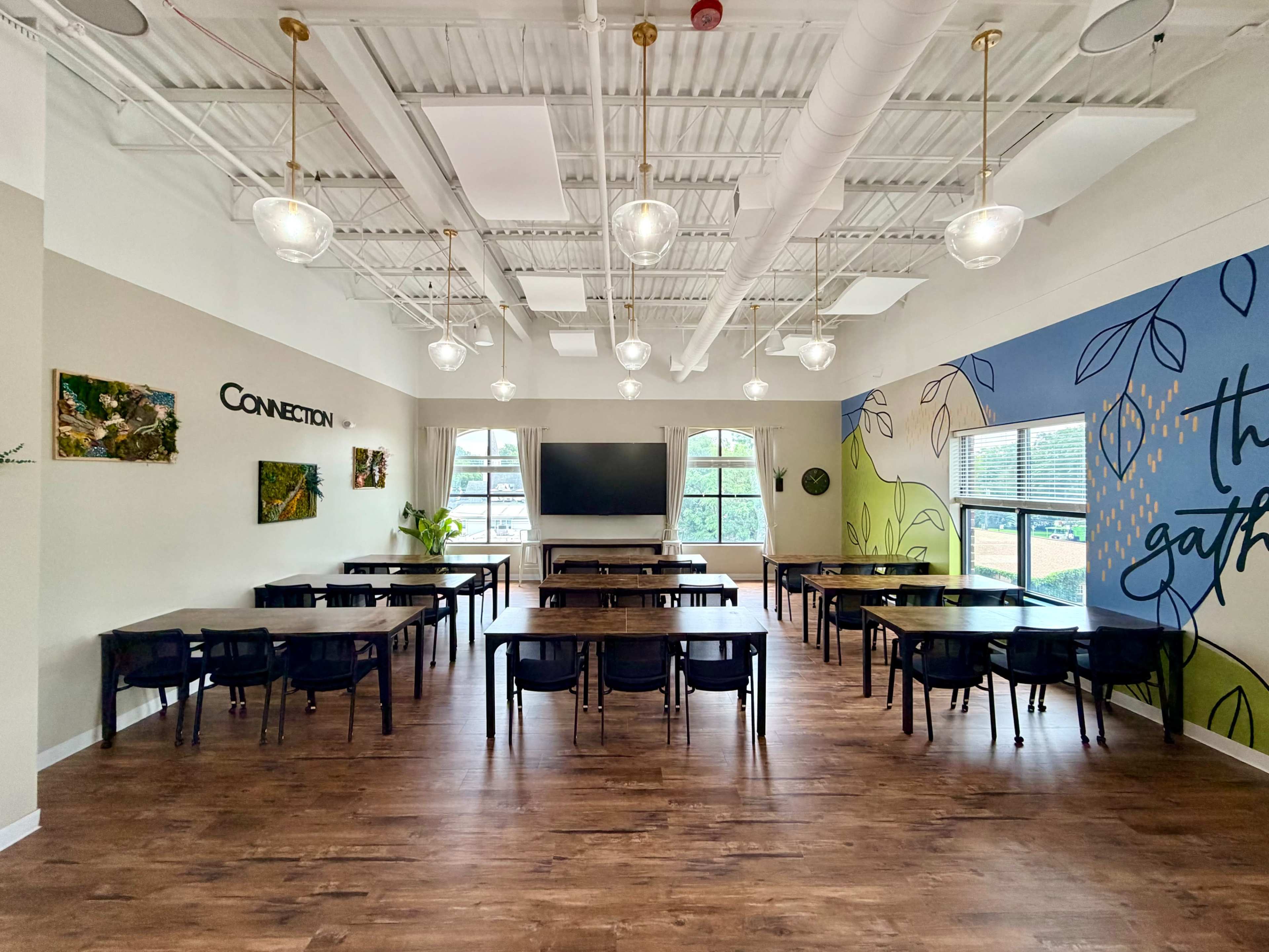 The image shows a spacious classroom with multiple long tables and chairs arranged in a neat layout, large windows allowing natural light, and a colorful mural on one wall.