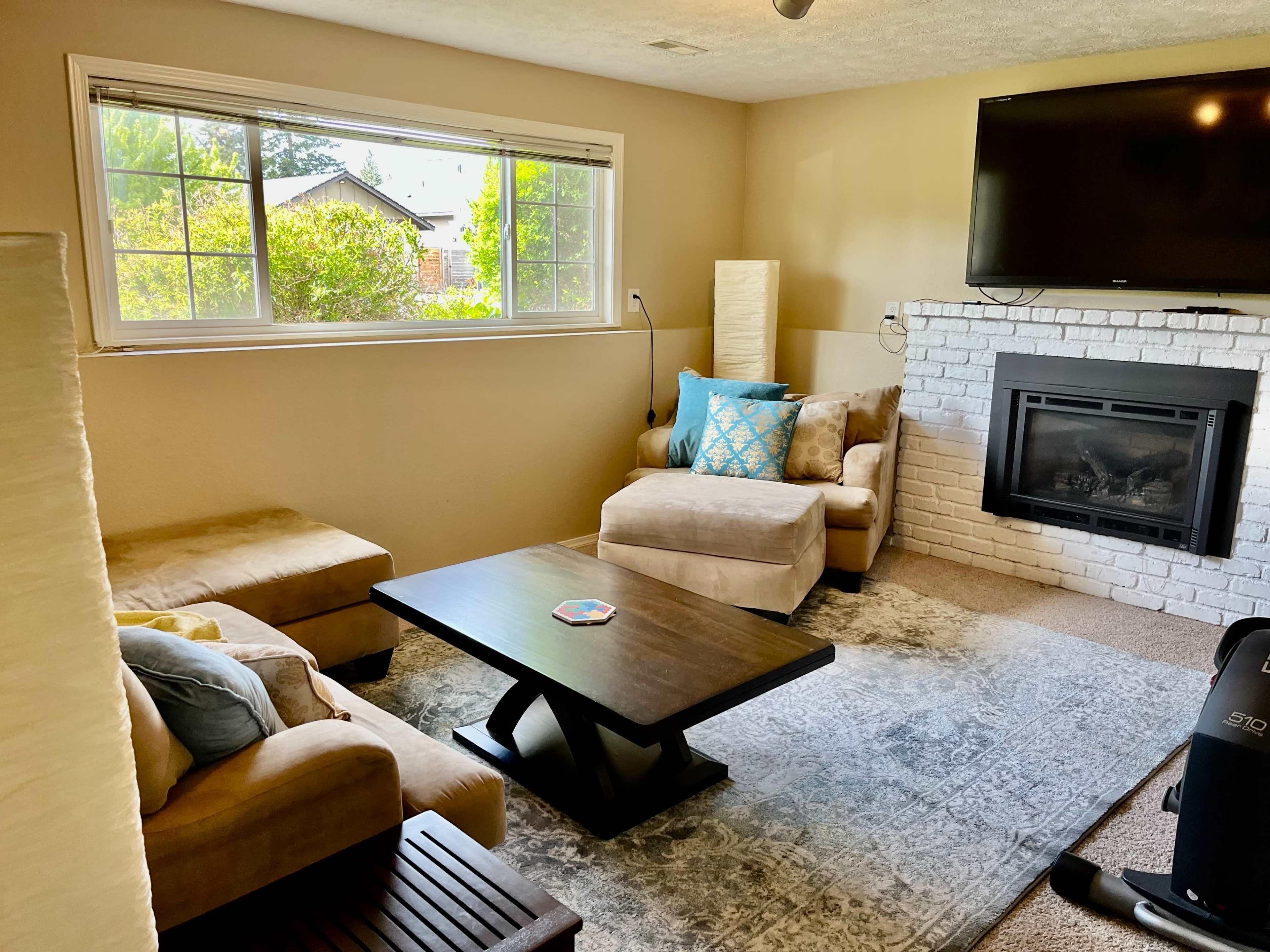 A cozy living room with a light-colored sofa, a chaise lounge, a coffee table, and a television above a white brick fireplace, illuminated by natural light from a large window.