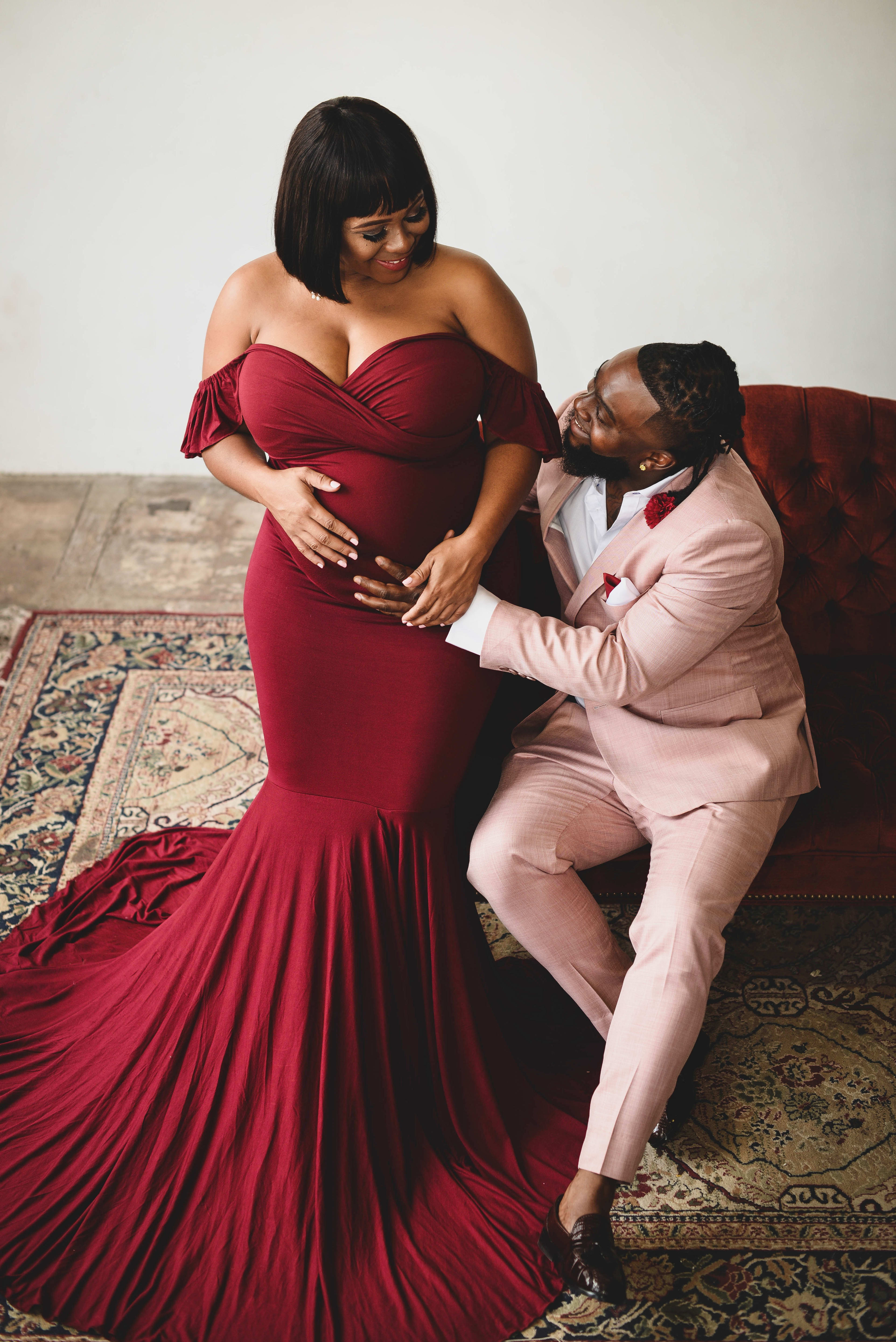 Una mujer embarazada y su pareja posando con un vestido rojo y un traje rosa para una sesión de fotos de maternidad.