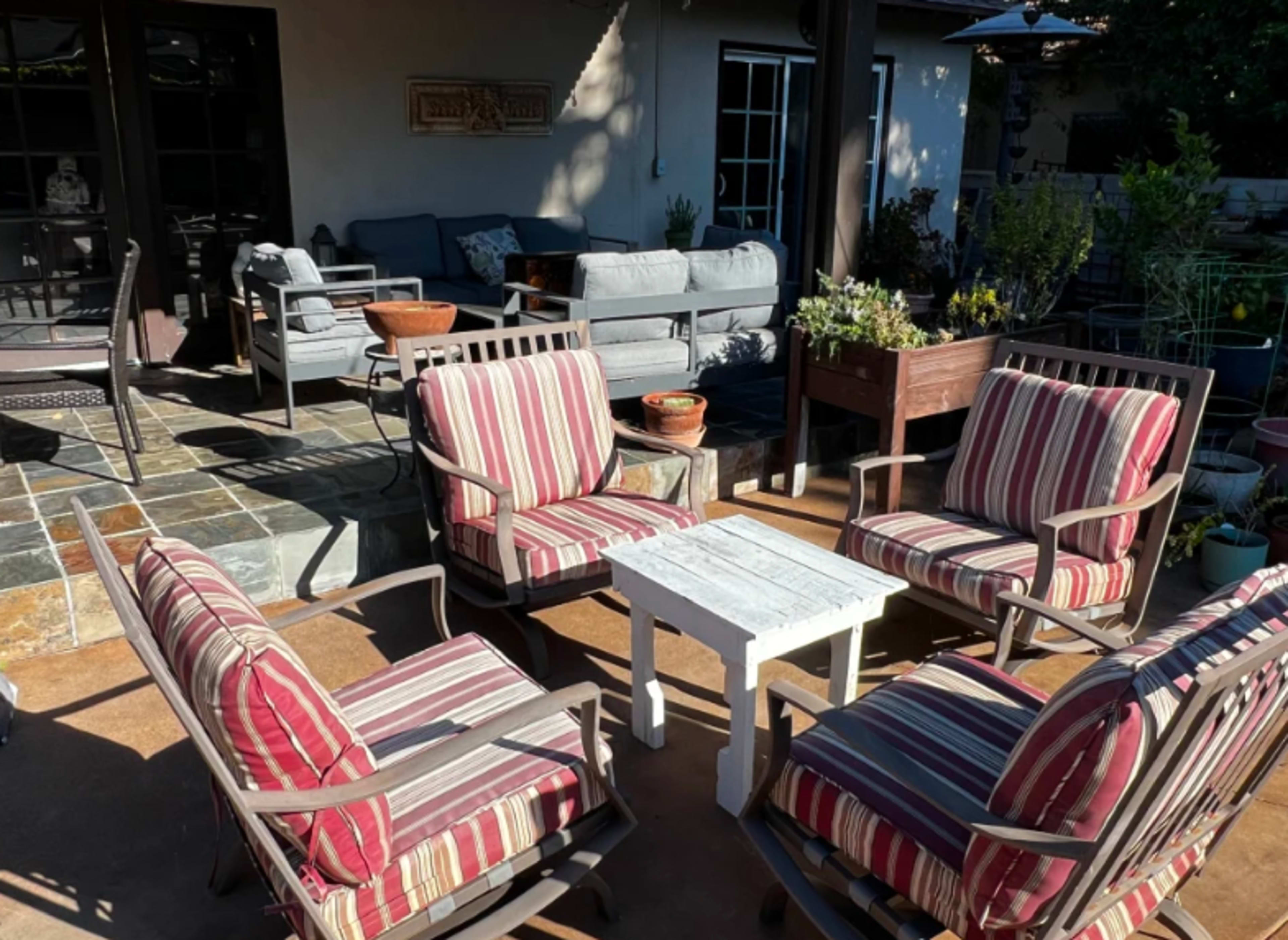 A patio area features striped armchairs arranged around a small white table, with potted plants and a shaded seating area in the background.