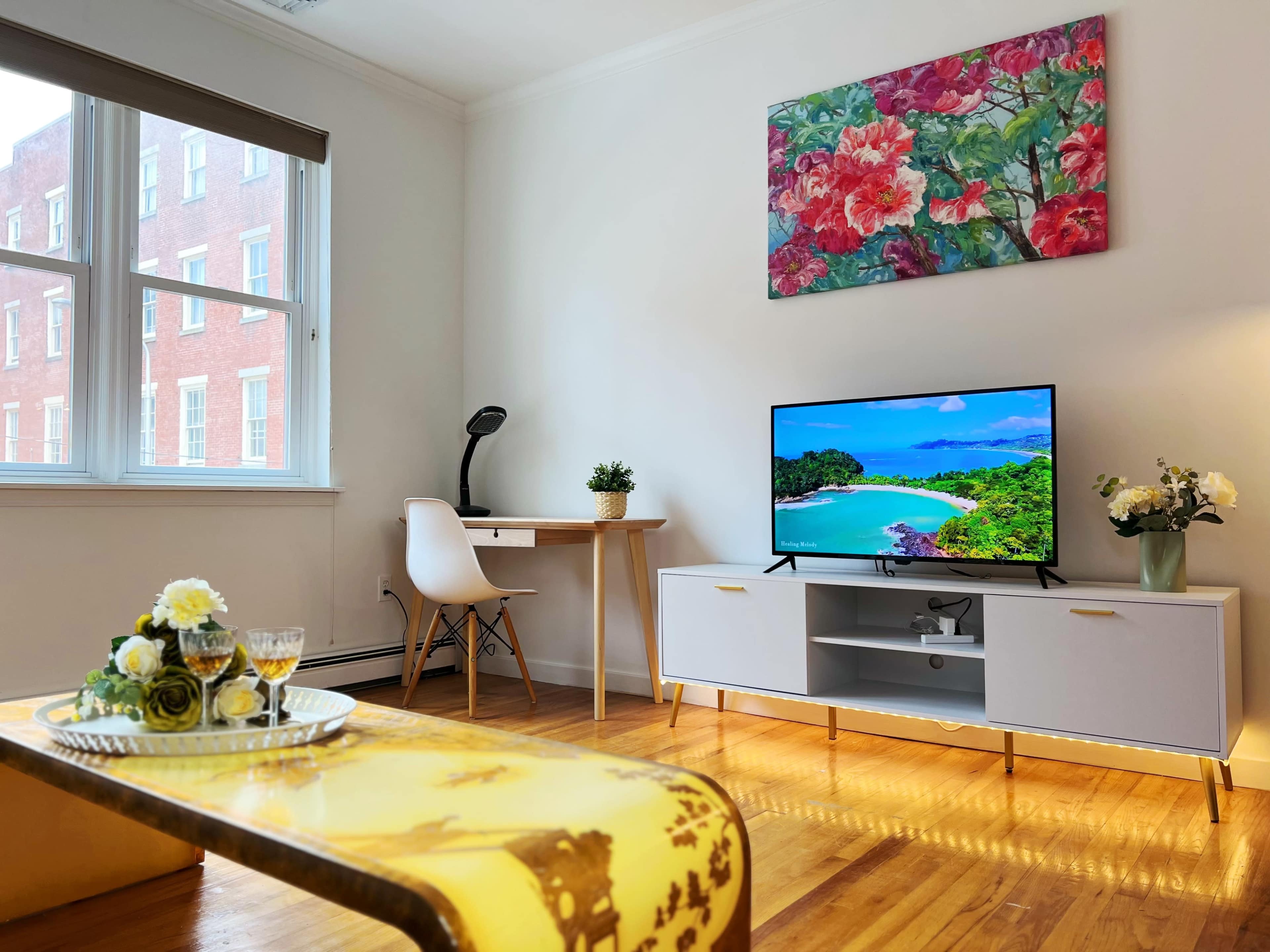 A bright living room featuring a television on a white cabinet, a desk with a lamp, and a vase of flowers on a coffee table.