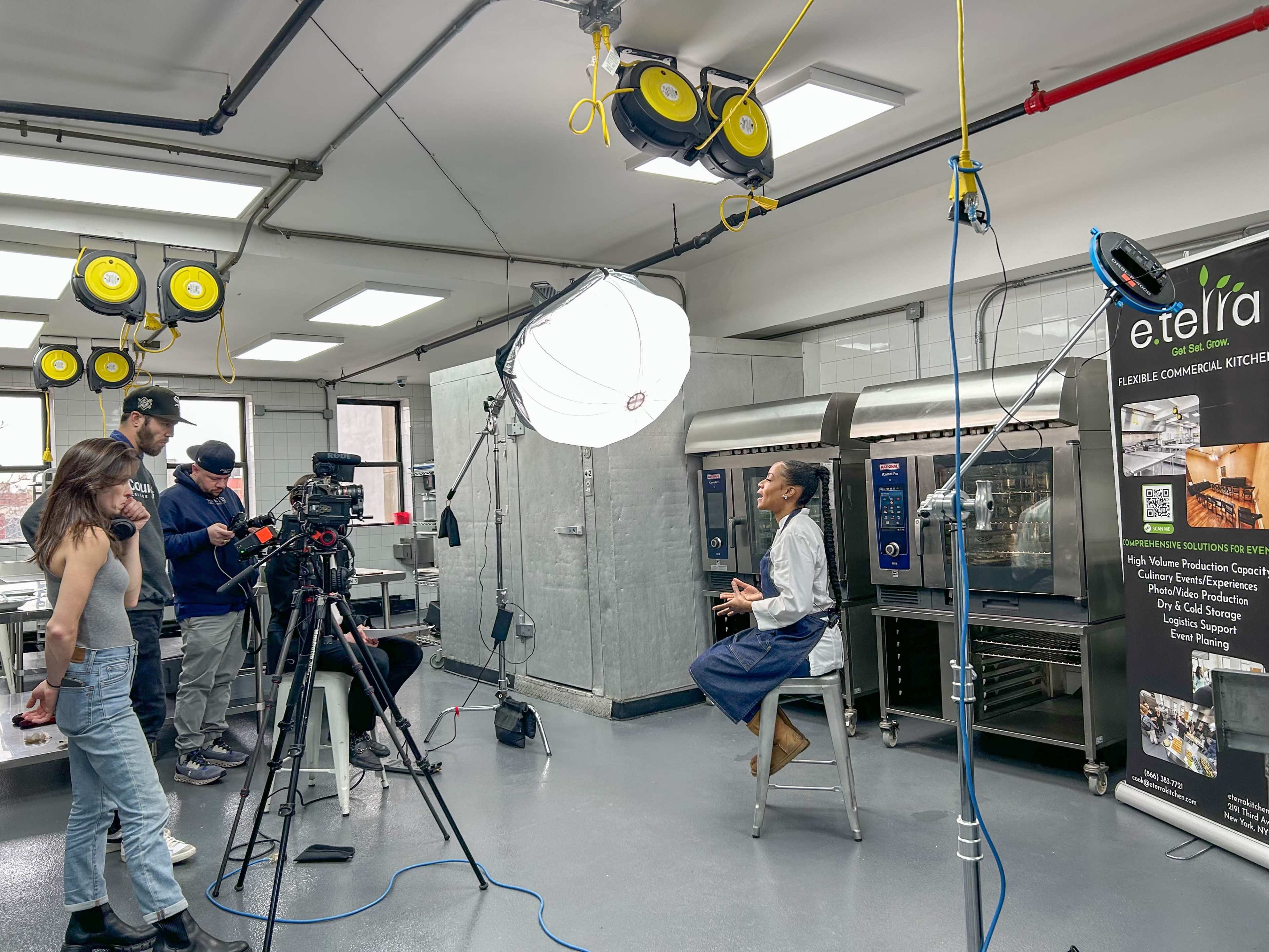 A person is seated for an interview in a commercial kitchen while a film crew sets up cameras and lights.