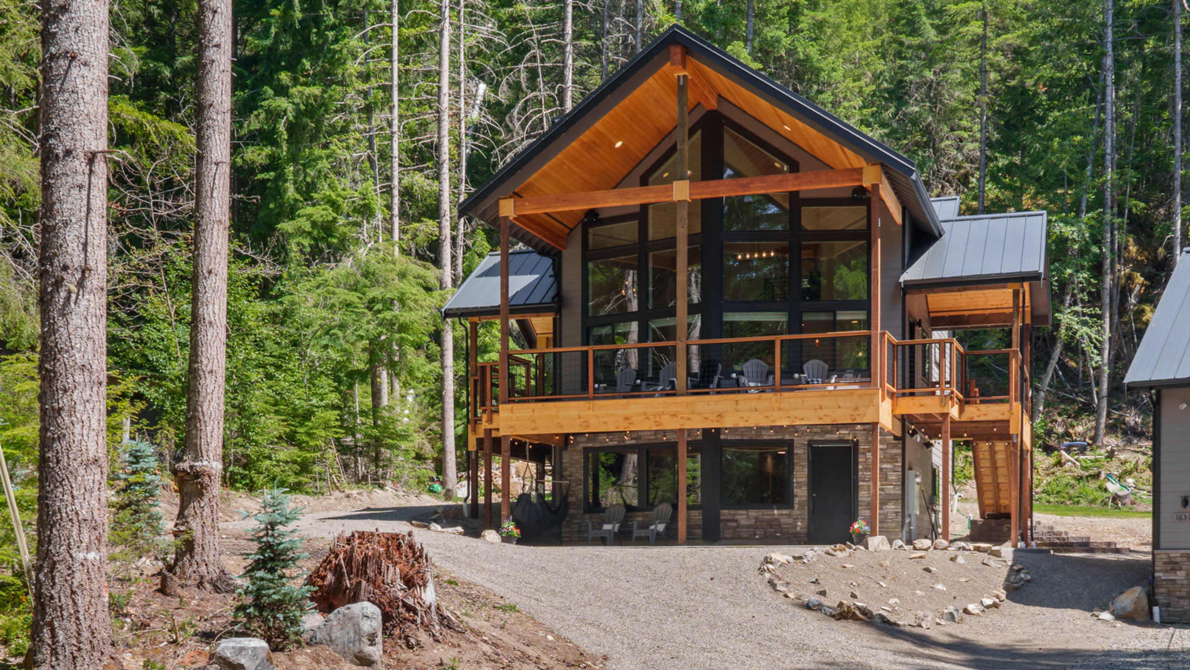 A modern cabin with large windows and a wooden deck is surrounded by tall pine trees and a gravel pathway.
