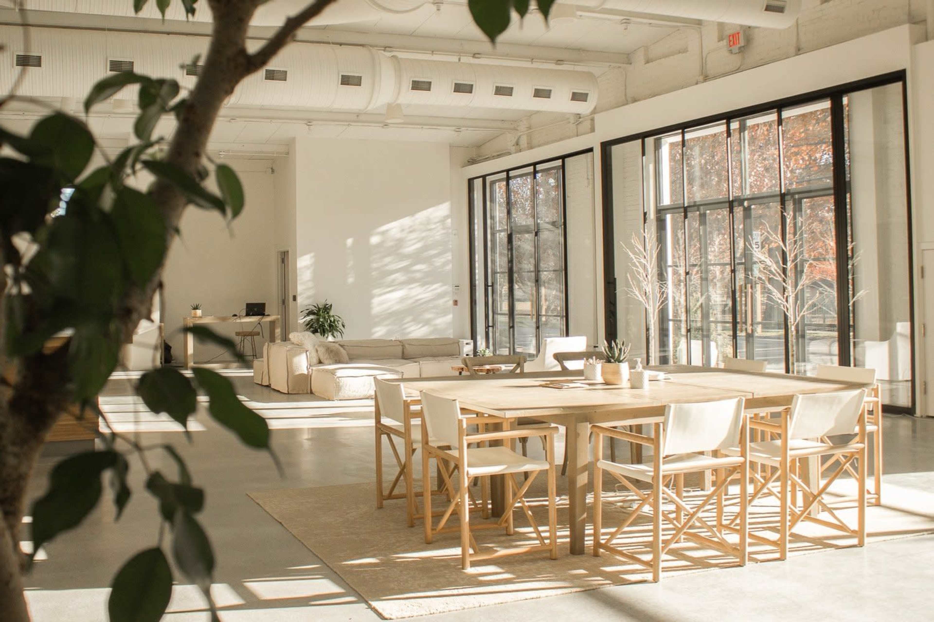 A spacious, modern interior features a large dining table with wooden chairs, a seating area, and expansive windows allowing natural light to fill the space.