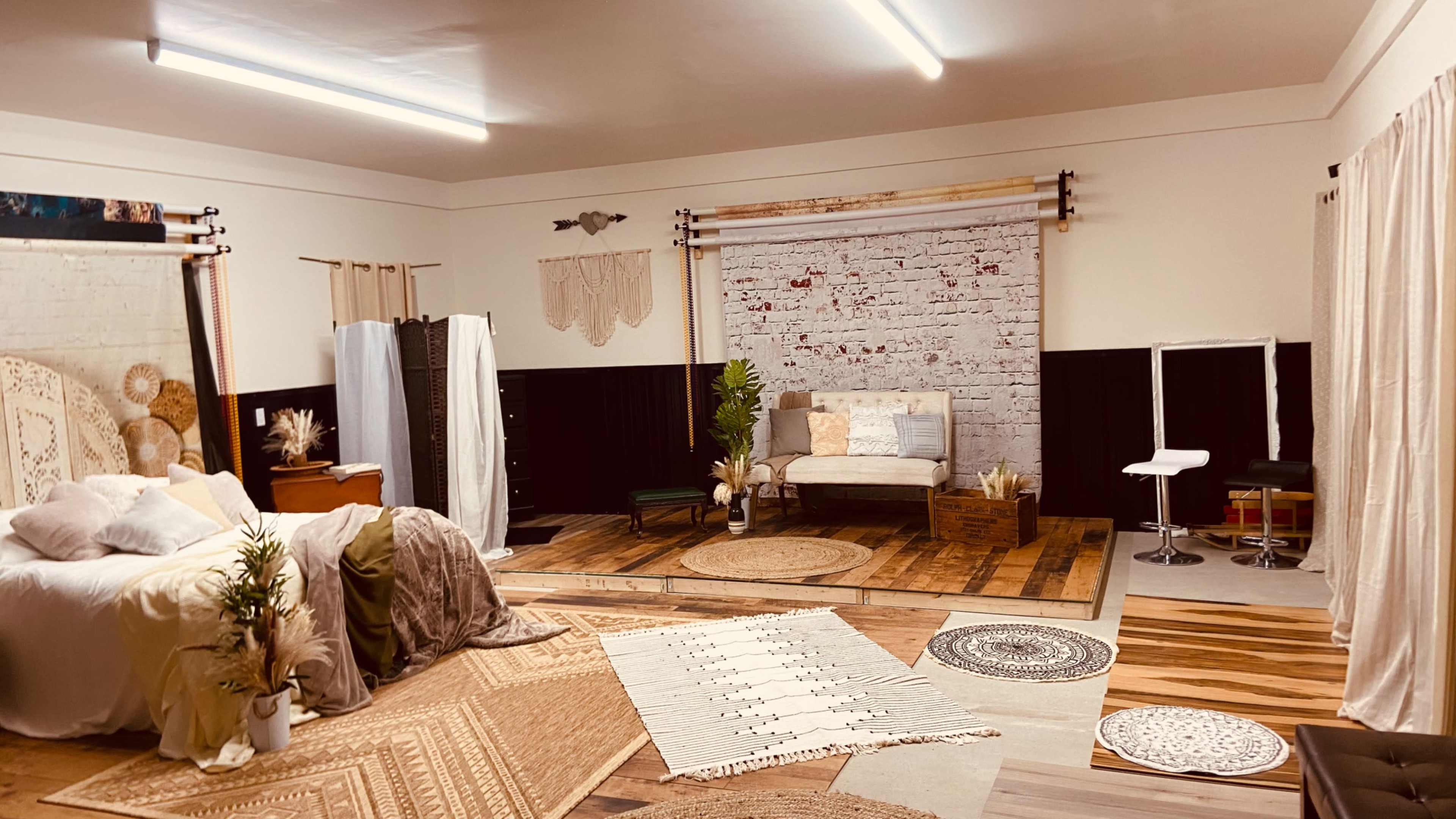 A spacious interior room designed for photography, featuring a bed, a couch, various rugs, and backdrop elements, with a mix of wood and fabric textures.
