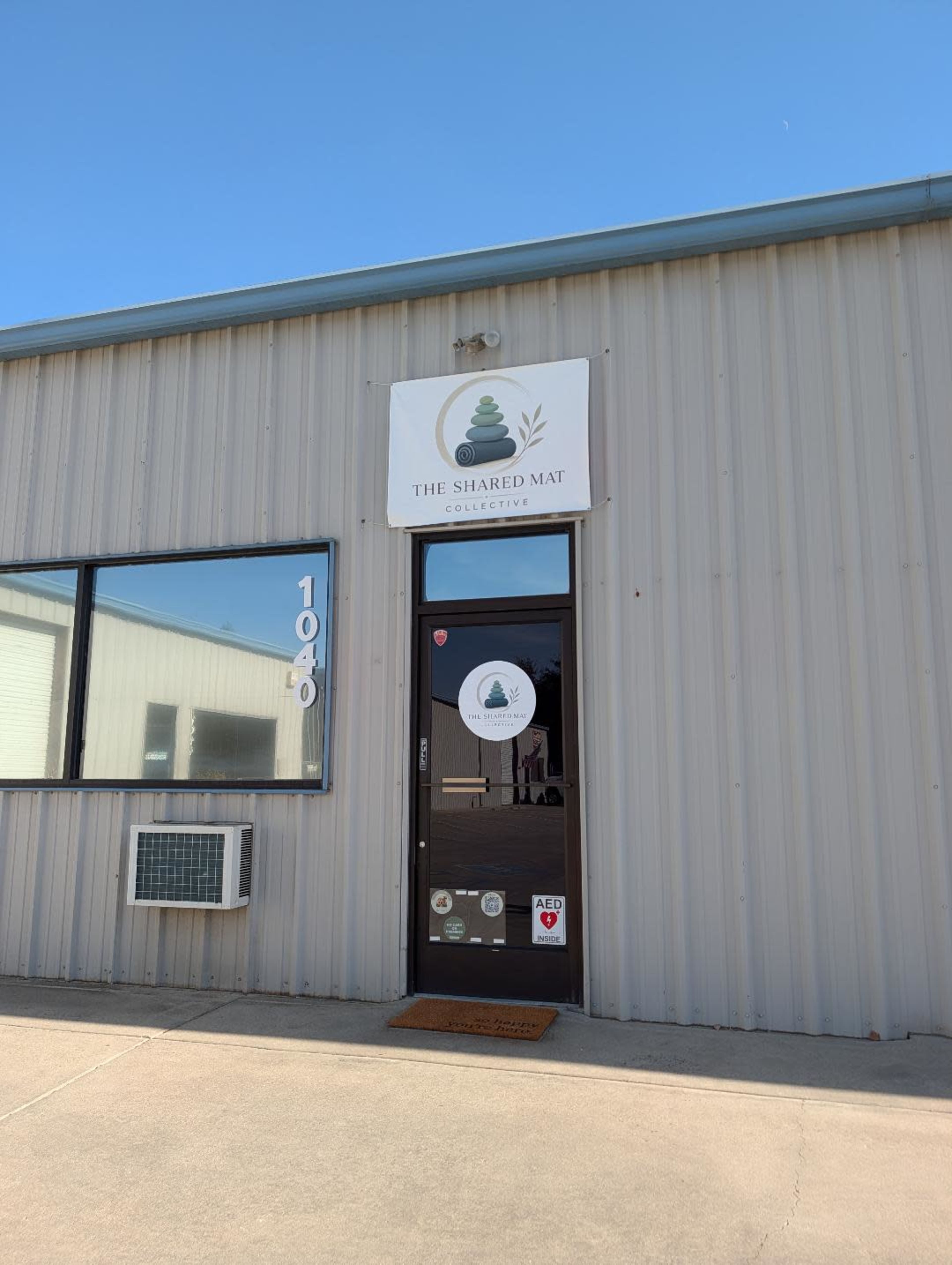 The image shows the entrance of a business called "The Shared Mat Collective," located in a metal-clad building with a clear blue sky above.