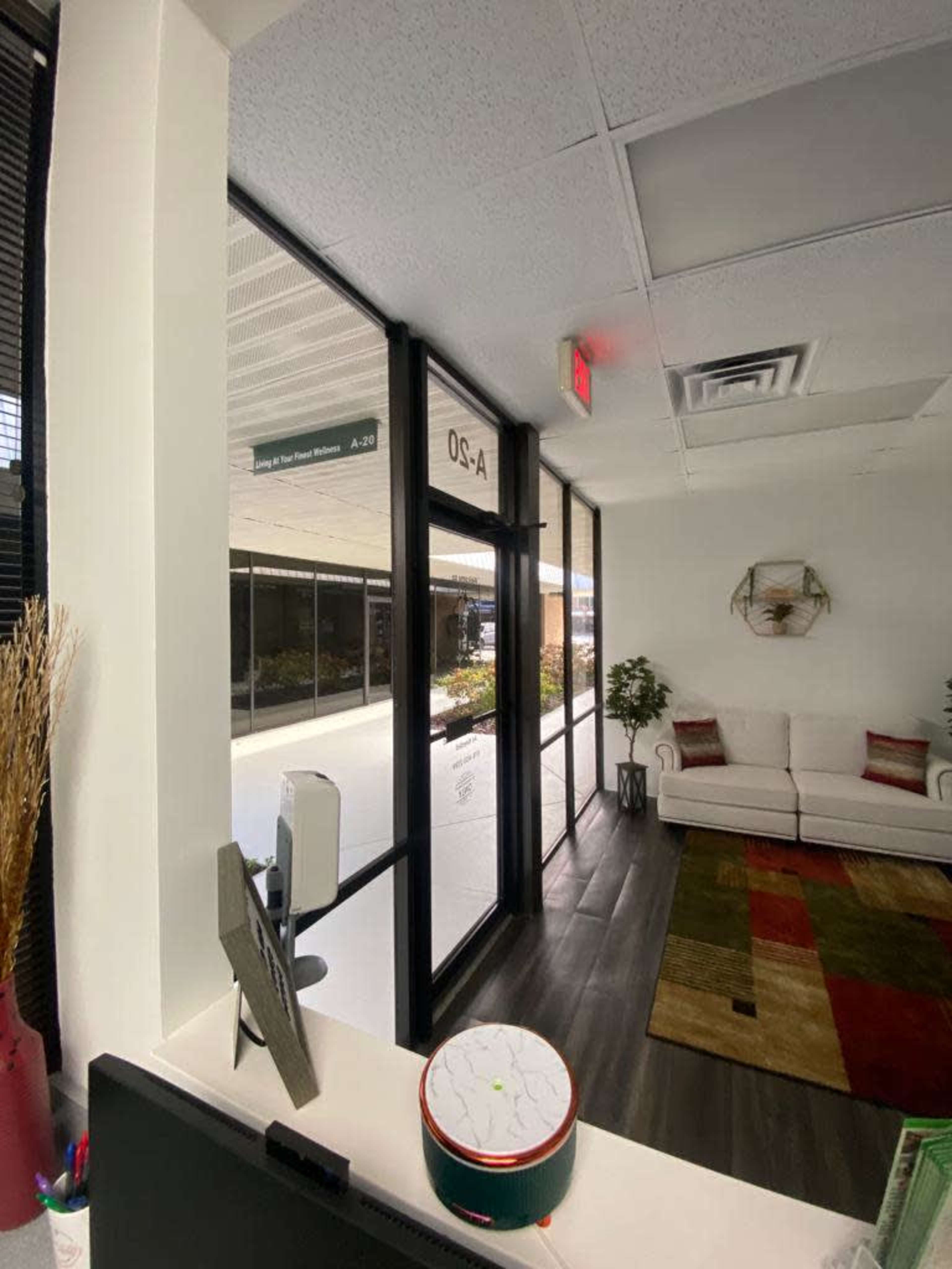 The image shows the interior of an office space with a reception area leading to a waiting room that features a couch and decorative plants.