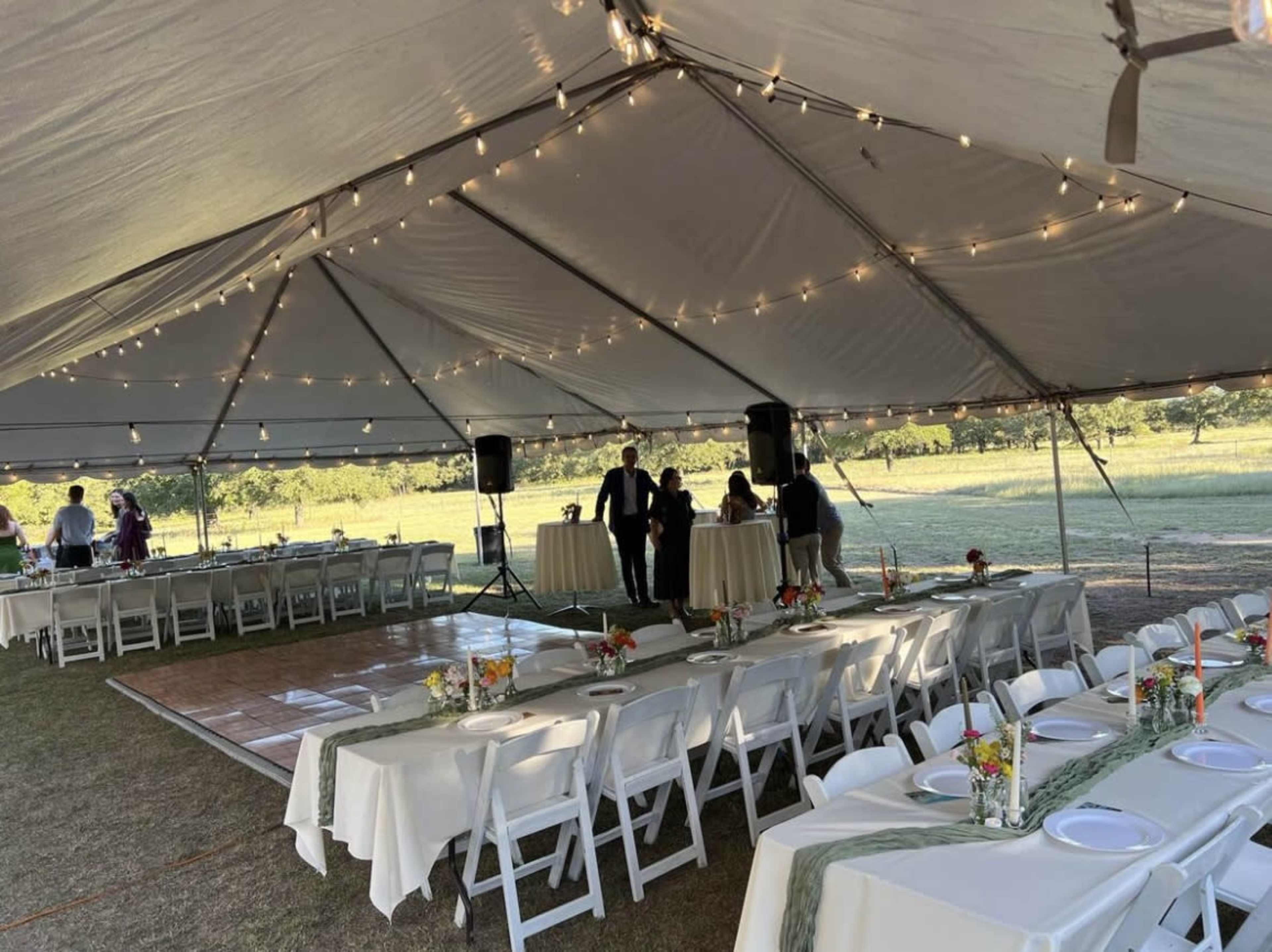 A large tent is set up with tables and chairs arranged for an outdoor event, featuring string lights overhead and a wooden dance floor.