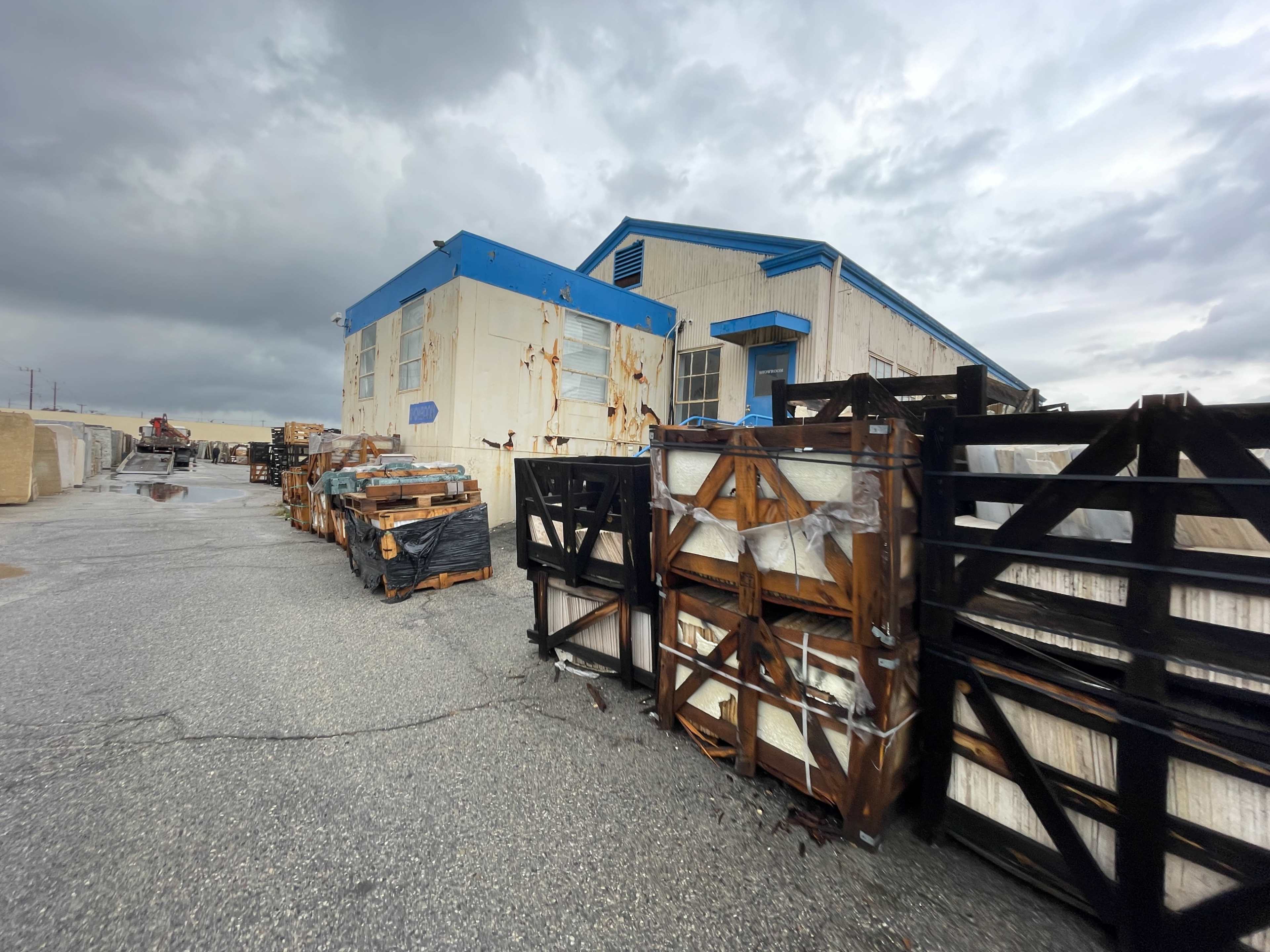 A weathered building with blue trim sits adjacent to stacks of wooden crates in a gravel area under a cloudy sky.
