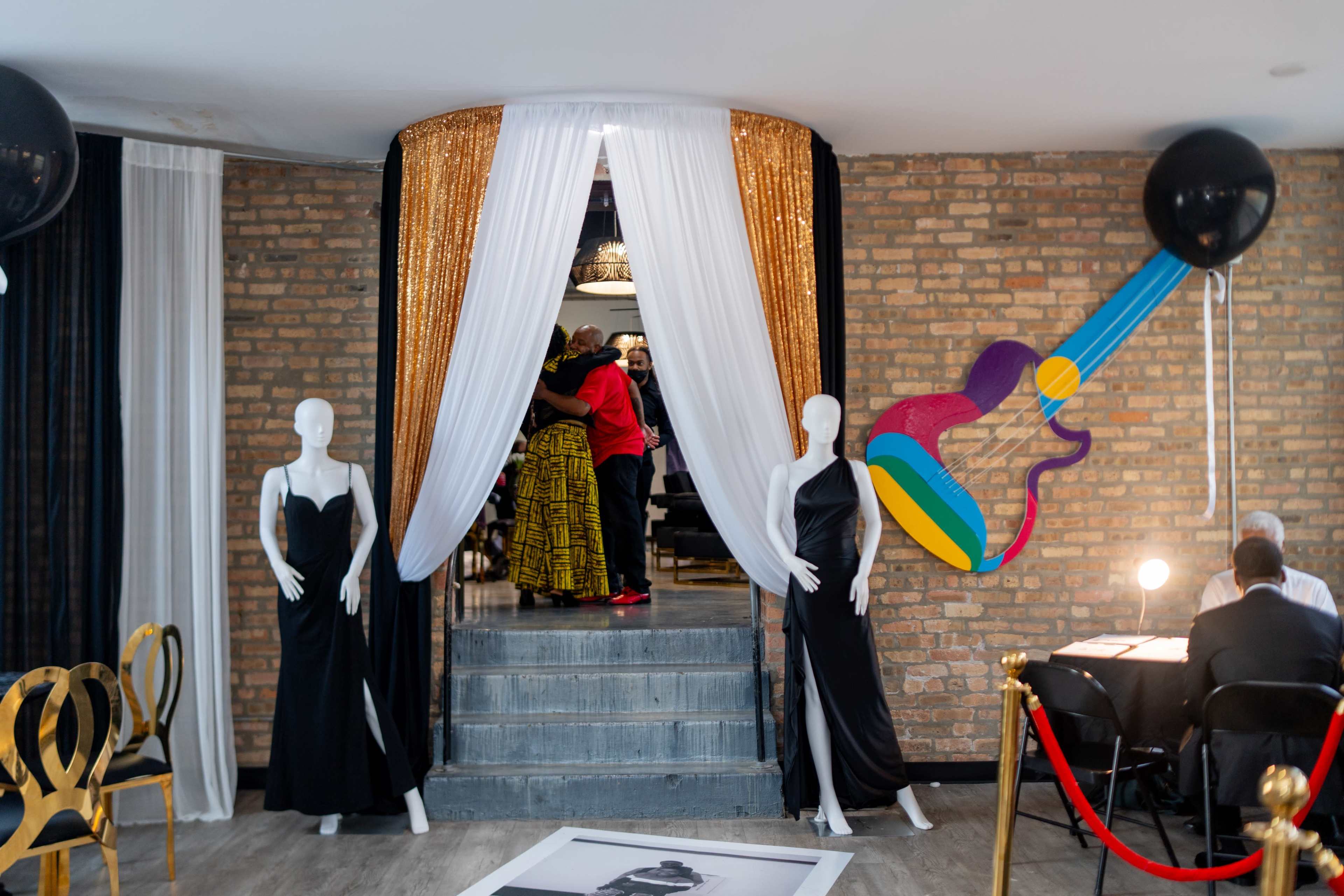 The image shows a stylish event space with mannequins dressed in black gowns, a staircase leading to a decorated area, and a colorful guitar mural on the wall.