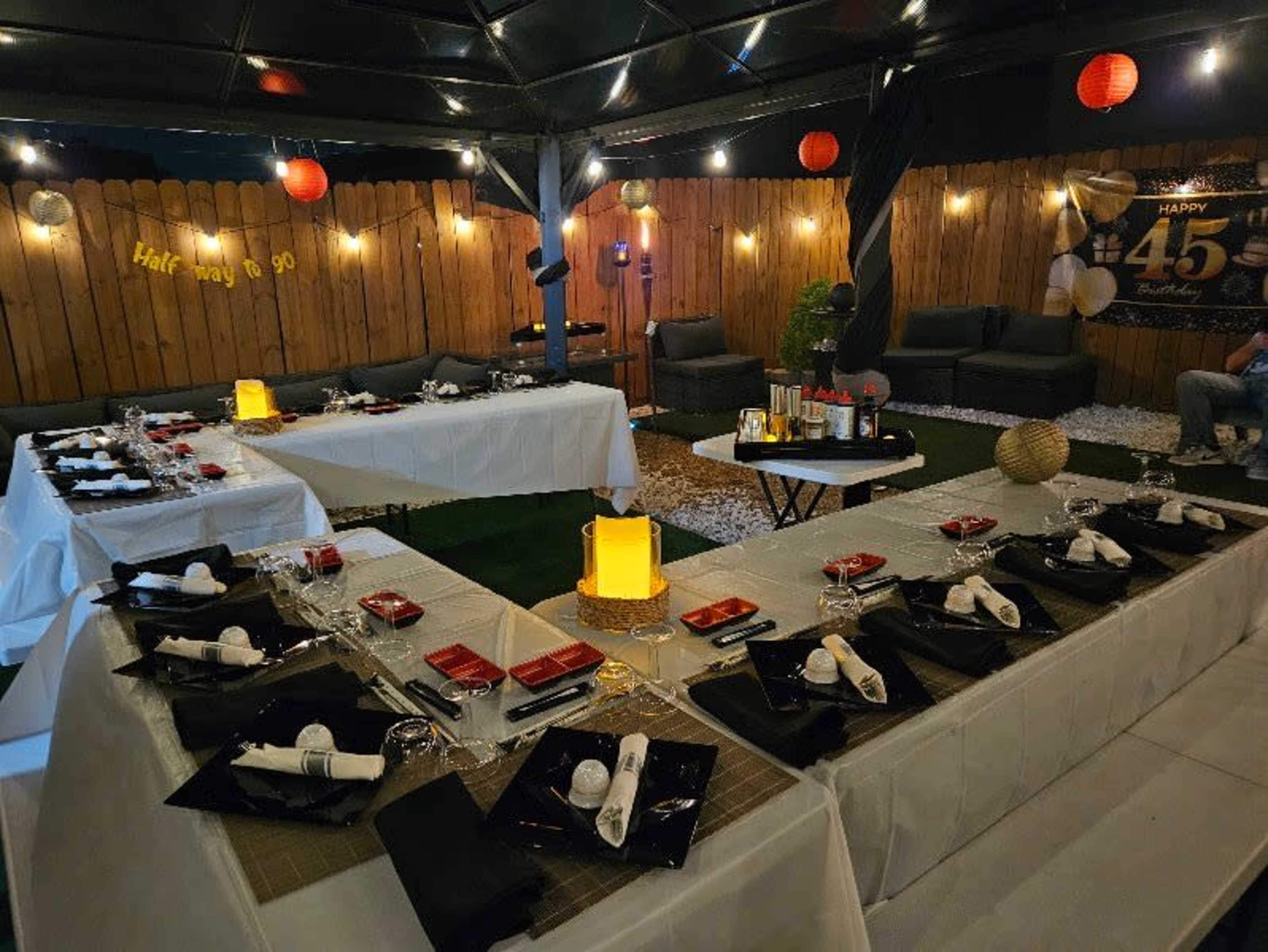 A canopy-covered outdoor space is set up for a dining event, featuring tables with white tablecloths, black napkins, and decorative centerpieces, illuminated by warm lights and lanterns.