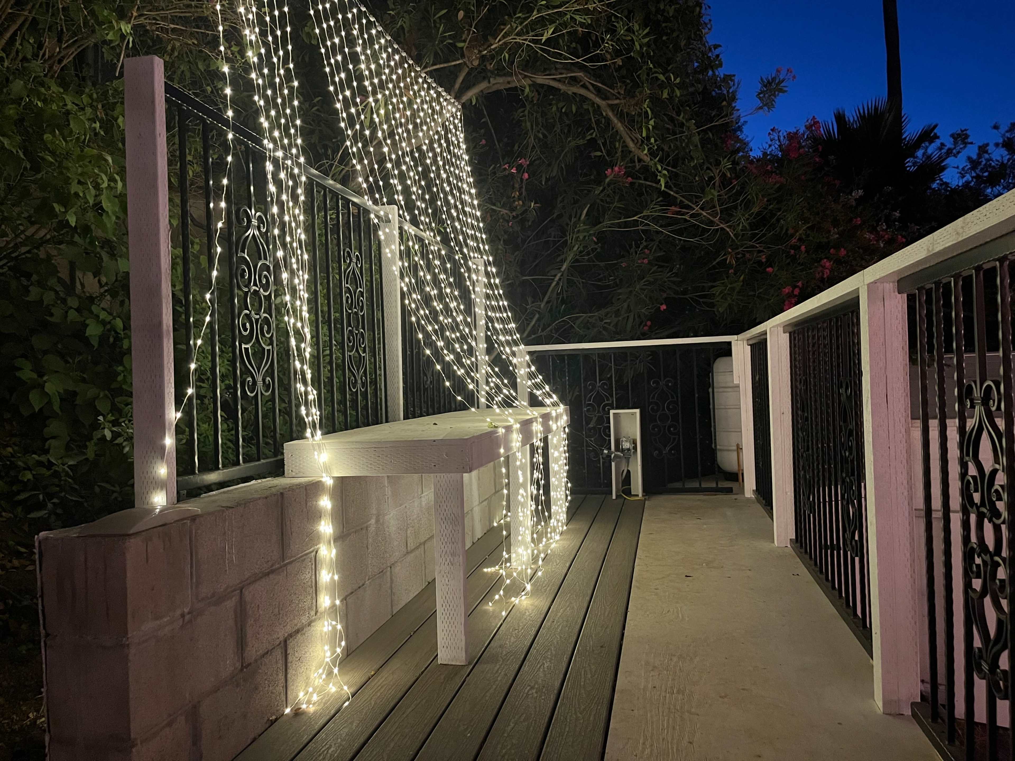 A patio area decorated with string lights hanging over a white bench against a backdrop of greenery during twilight.