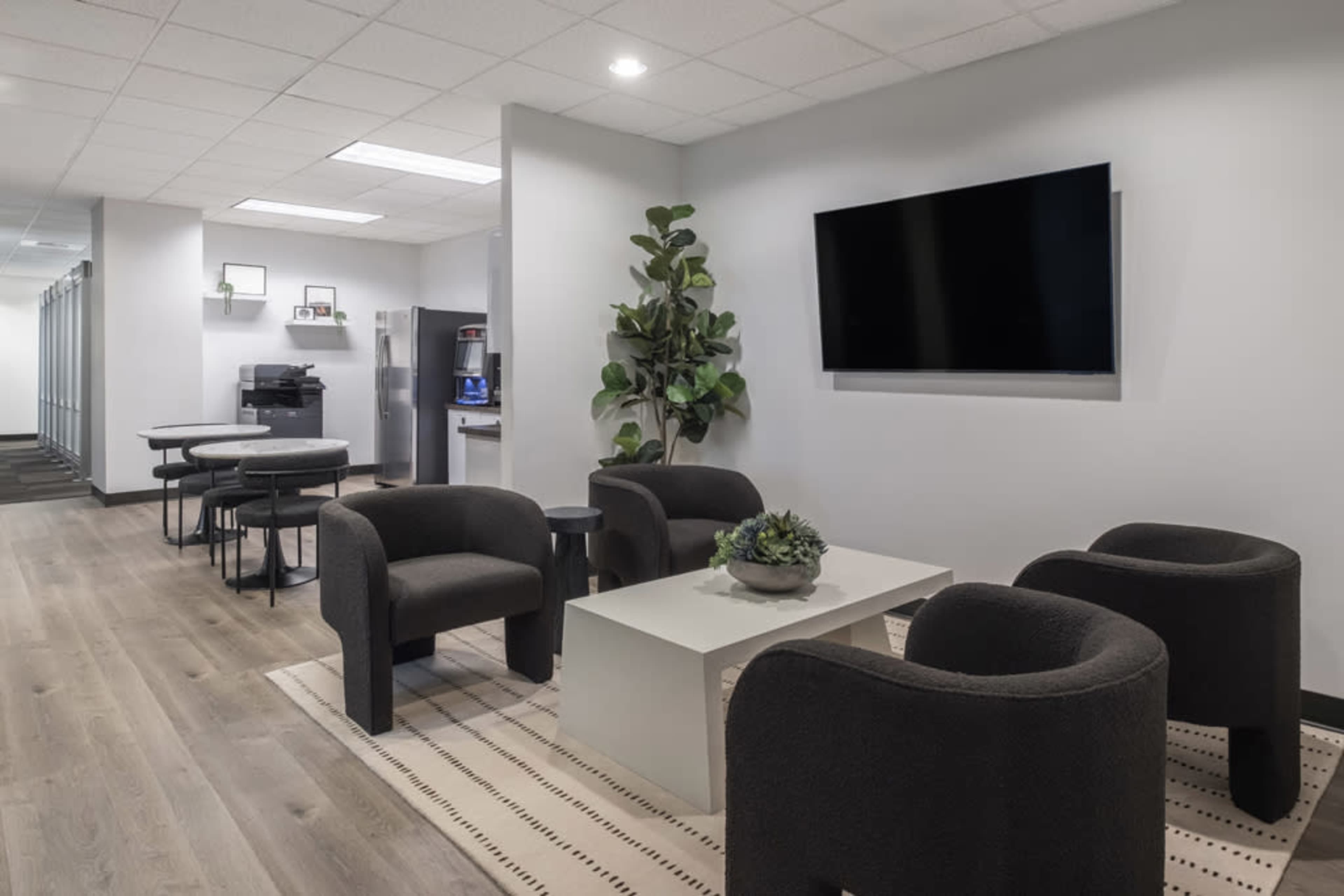 The image shows a modern office lounge area with four chairs, a coffee table, and a wall-mounted television.