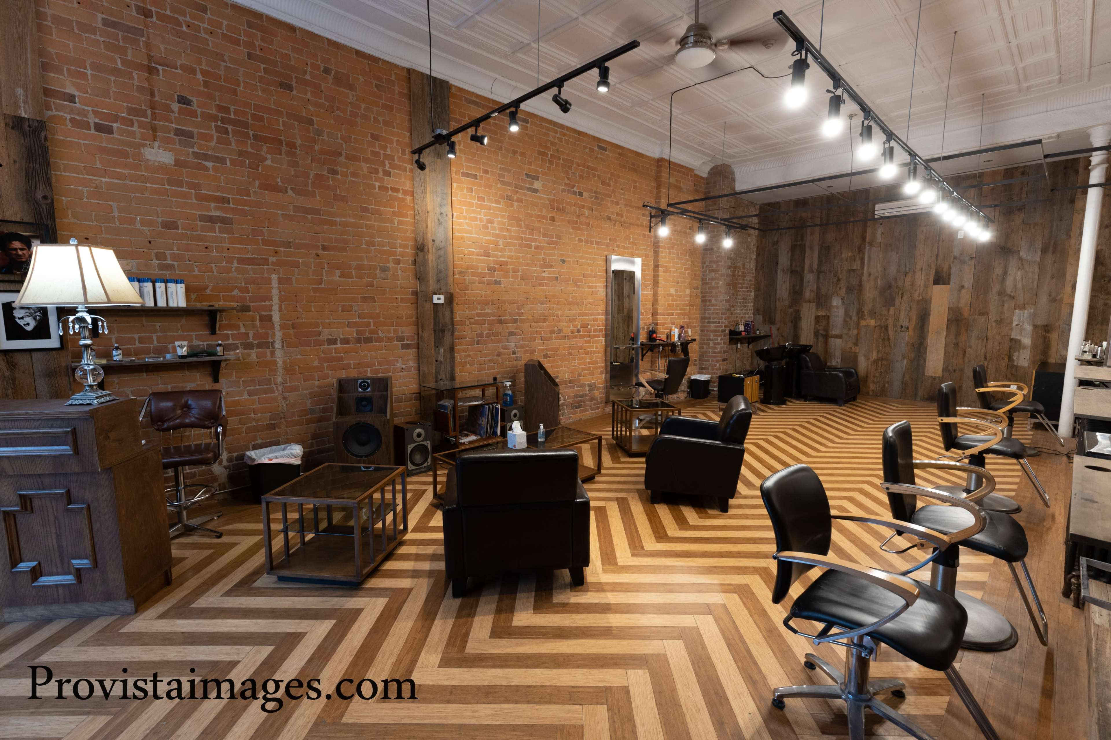 A spacious barber shop with a brick wall, wooden flooring arranged in a herringbone pattern, and modern black salon chairs.