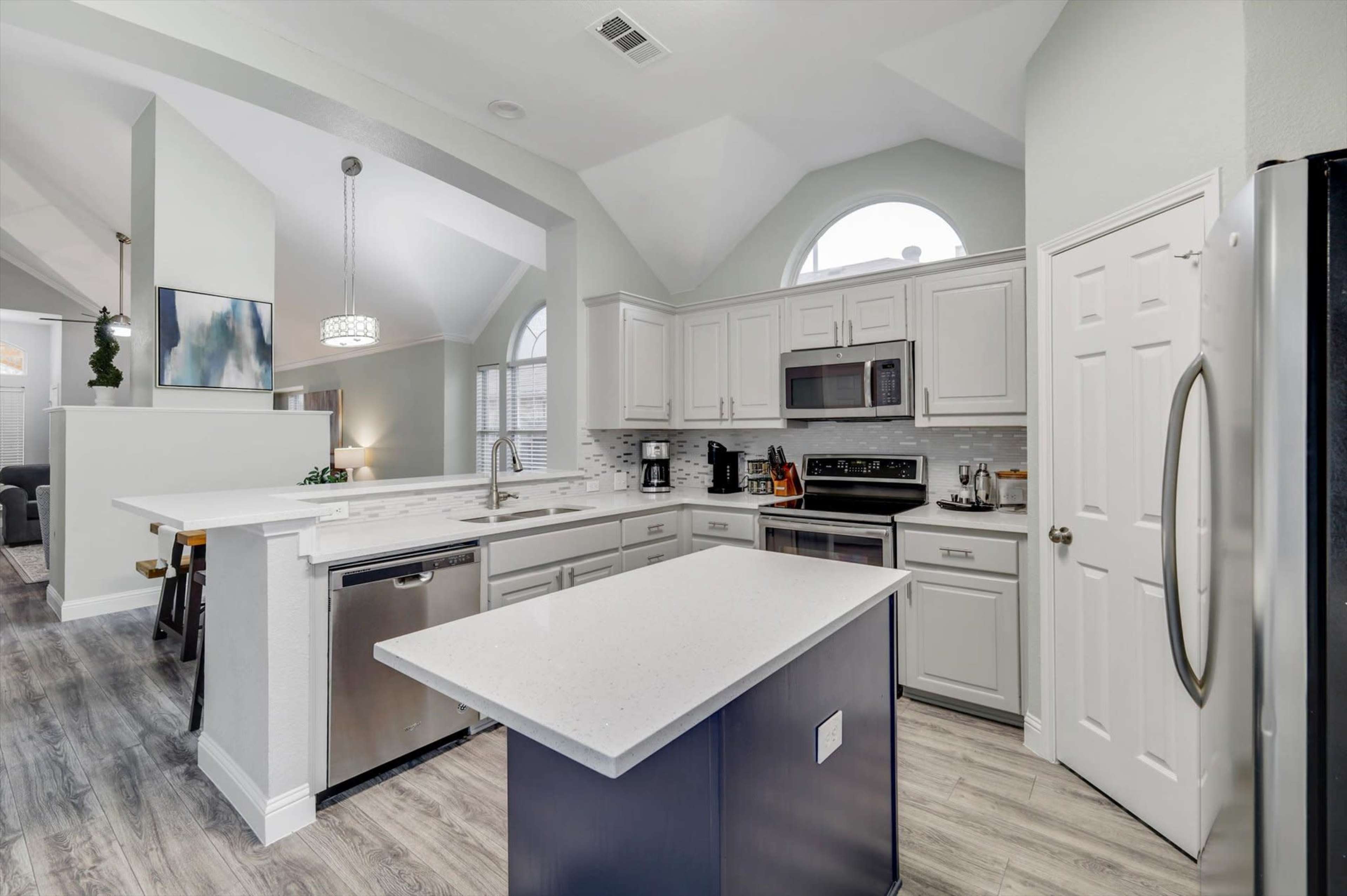 The image shows a modern kitchen featuring white cabinetry, stainless steel appliances, and a central island with a dark blue base.