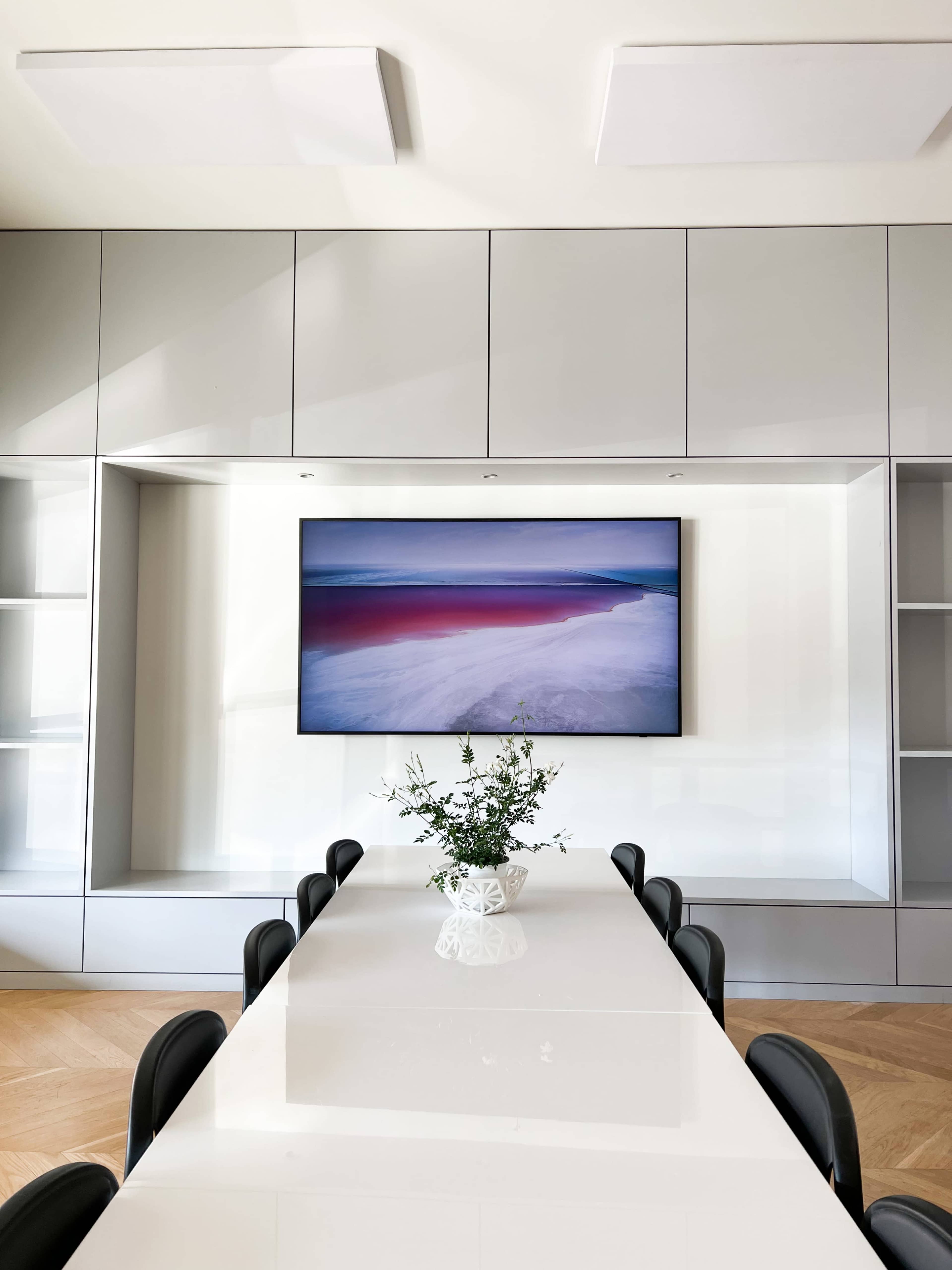 A modern conference room features a long white table with black chairs and a television mounted on a sleek gray cabinetry wall.