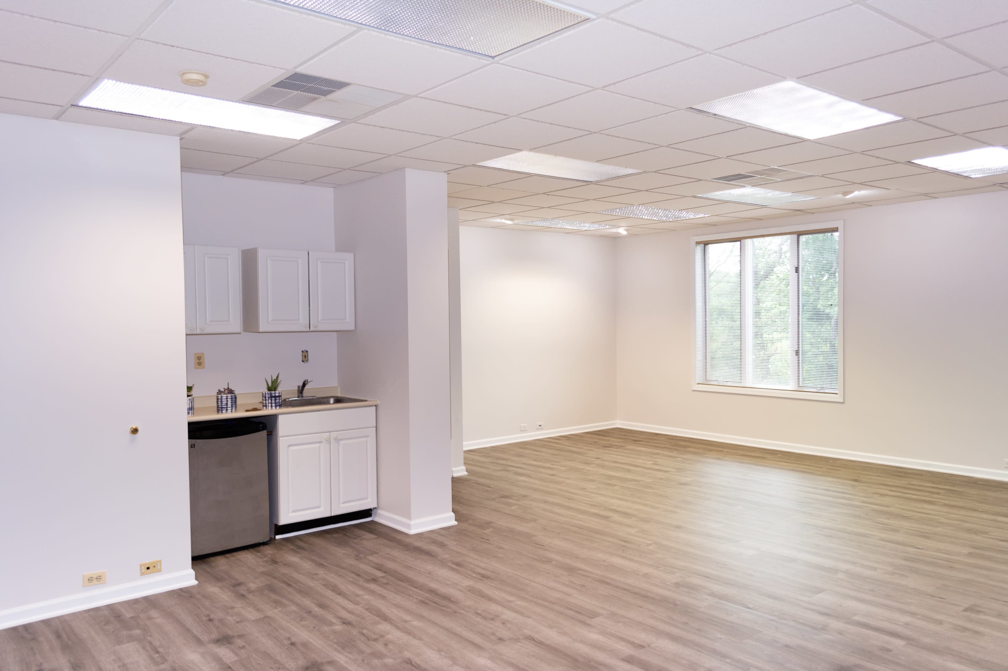 A spacious, empty room with light-colored walls, a small kitchenette in one corner, and a large window allowing natural light.