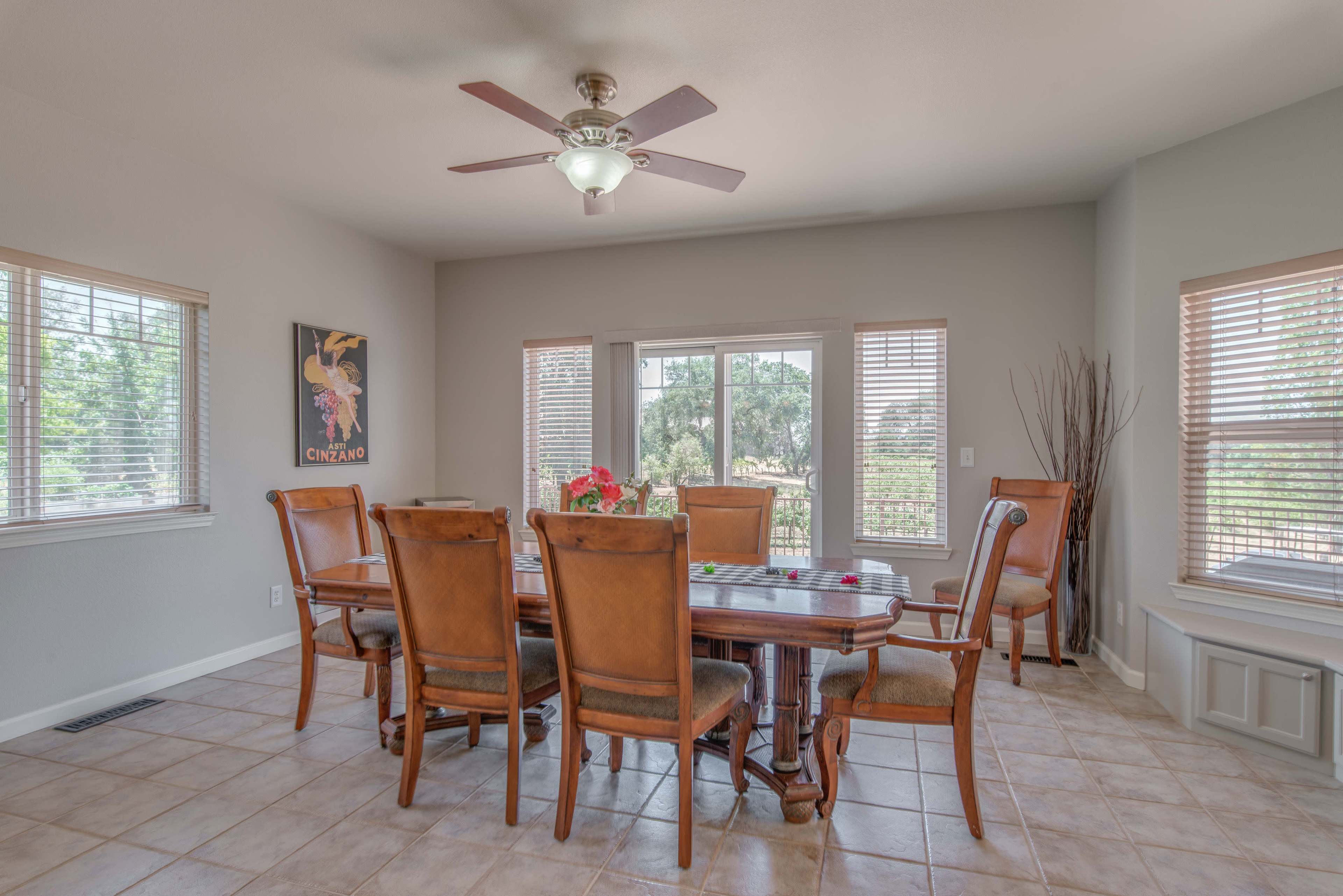 A dining room features a large wooden table surrounded by upholstered chairs, with a ceiling fan overhead and windows allowing natural light.