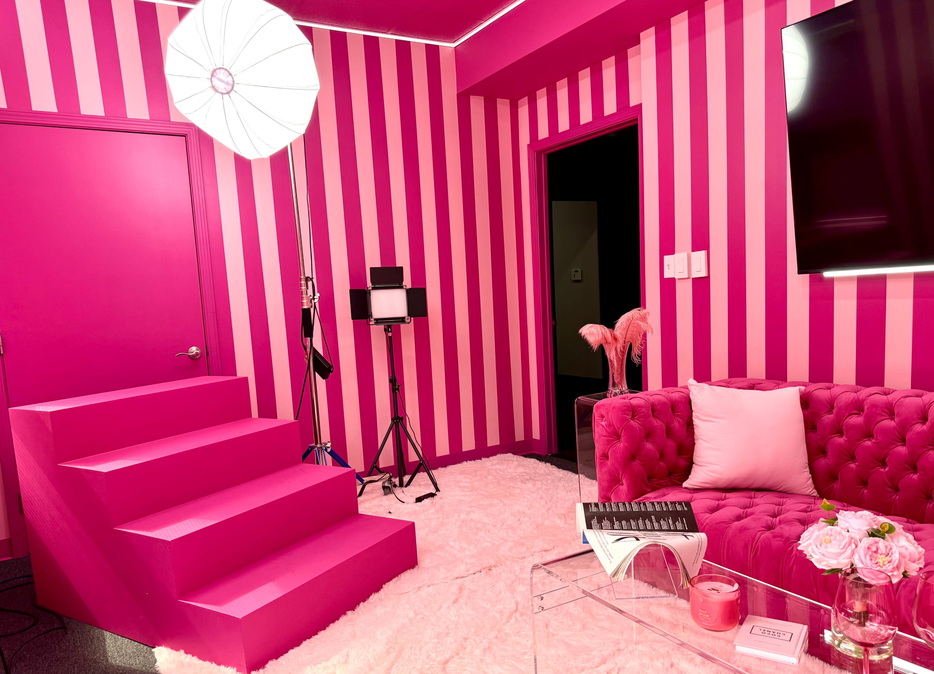 The image features a vibrant pink room with striped walls, a plush pink couch, a white rug, and a set of stairs leading to darker interior spaces.