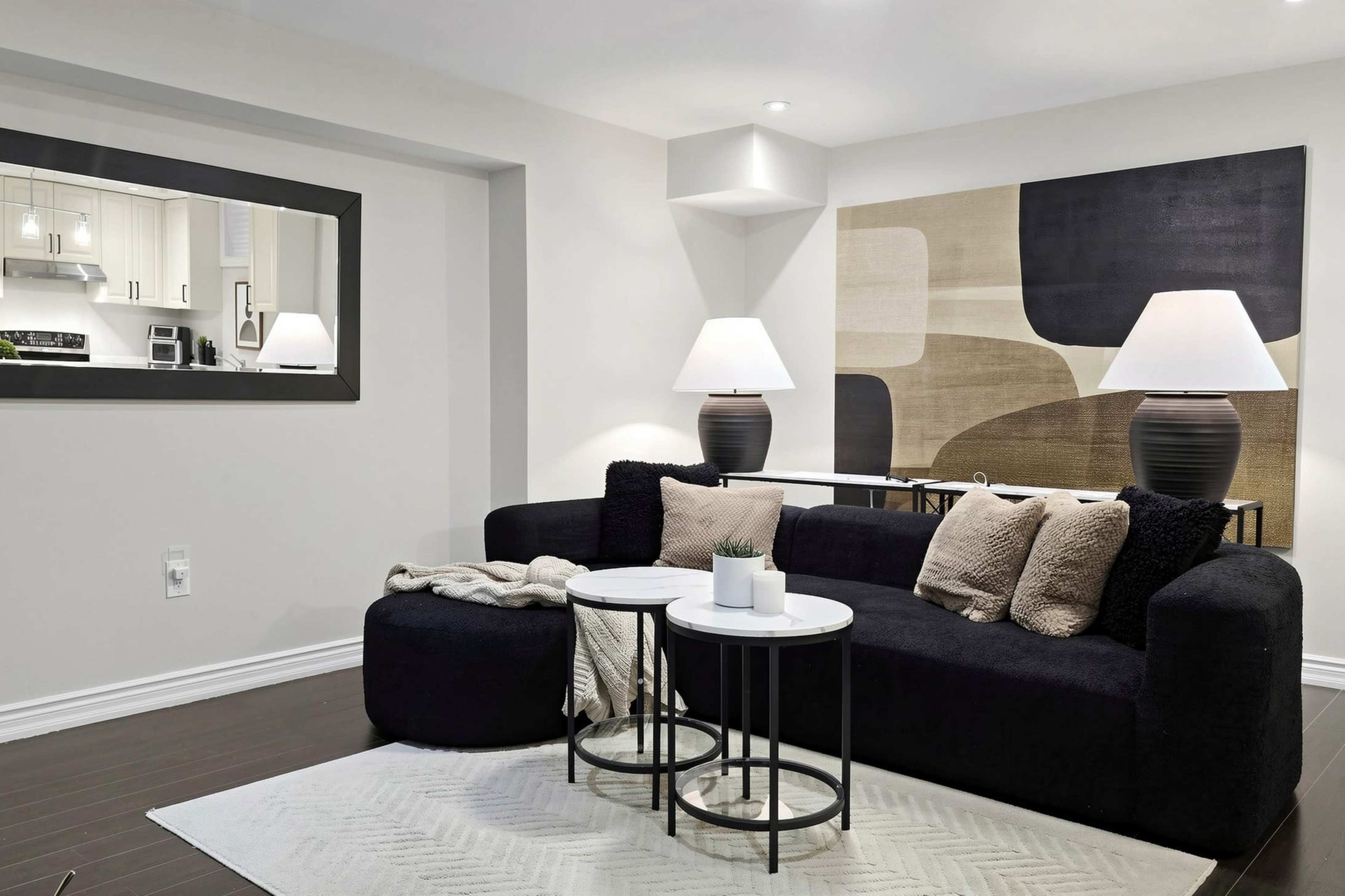 The scene depicts a modern living room featuring a black sectional sofa, two round side tables, and abstract wall art, illuminated by two table lamps.