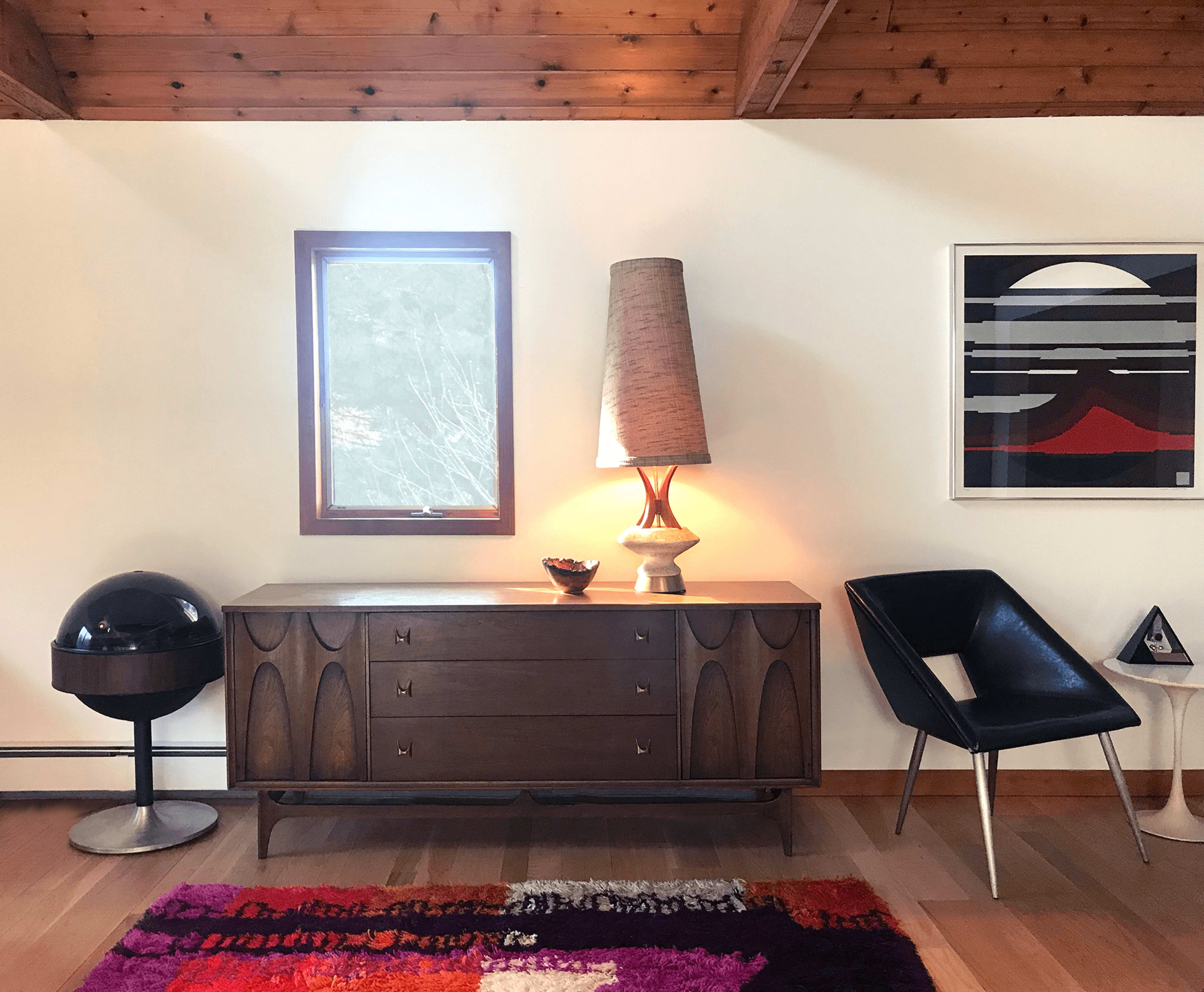 The image features a mid-century modern interior with a wooden dresser, a tall lamp, a round black chair, and a colorful rug against a wall with a window and framed artwork.