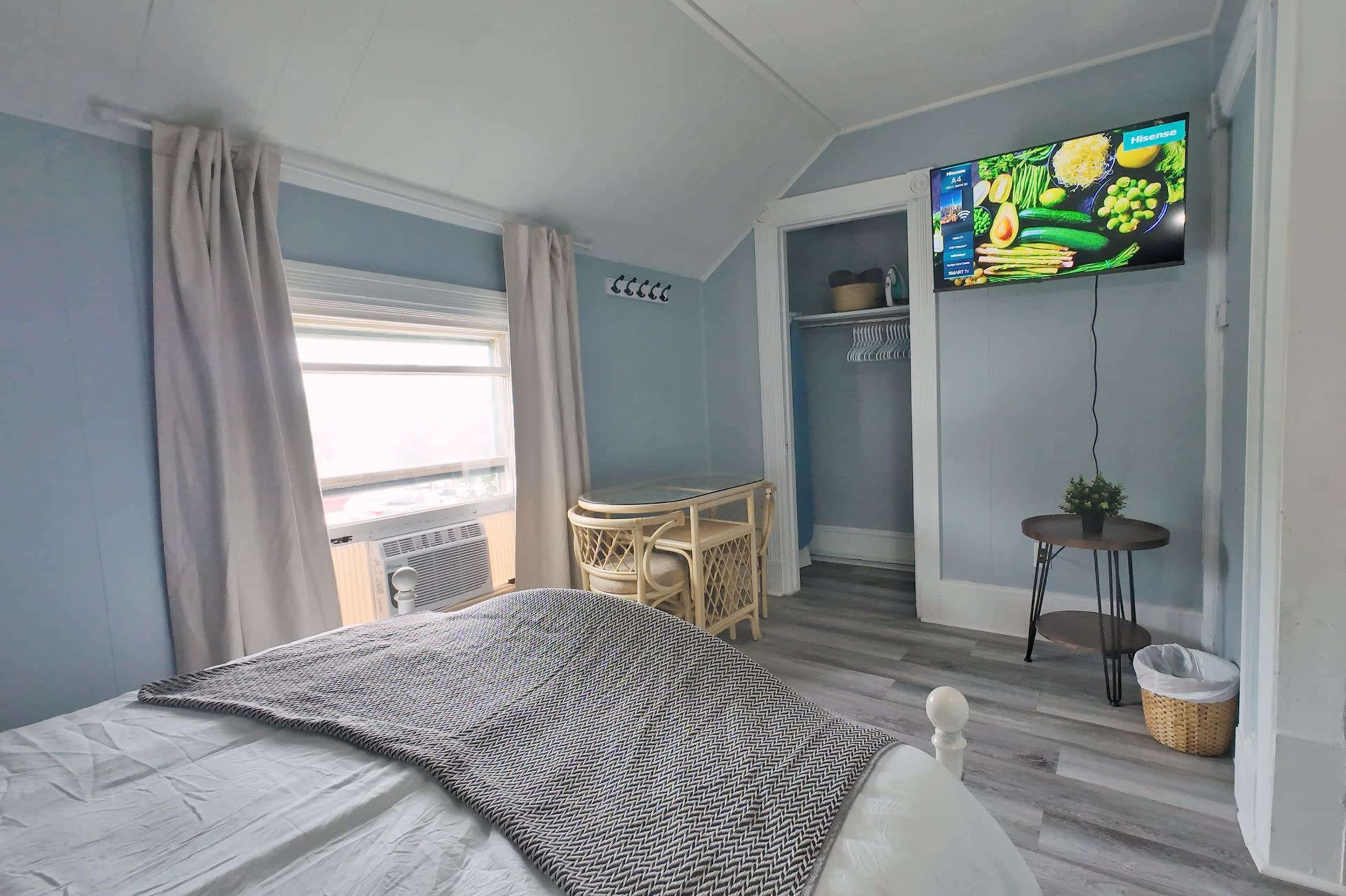 The image shows a cozy bedroom with a large window, a bed with a light-colored blanket, a small table, and a television mounted on the wall.