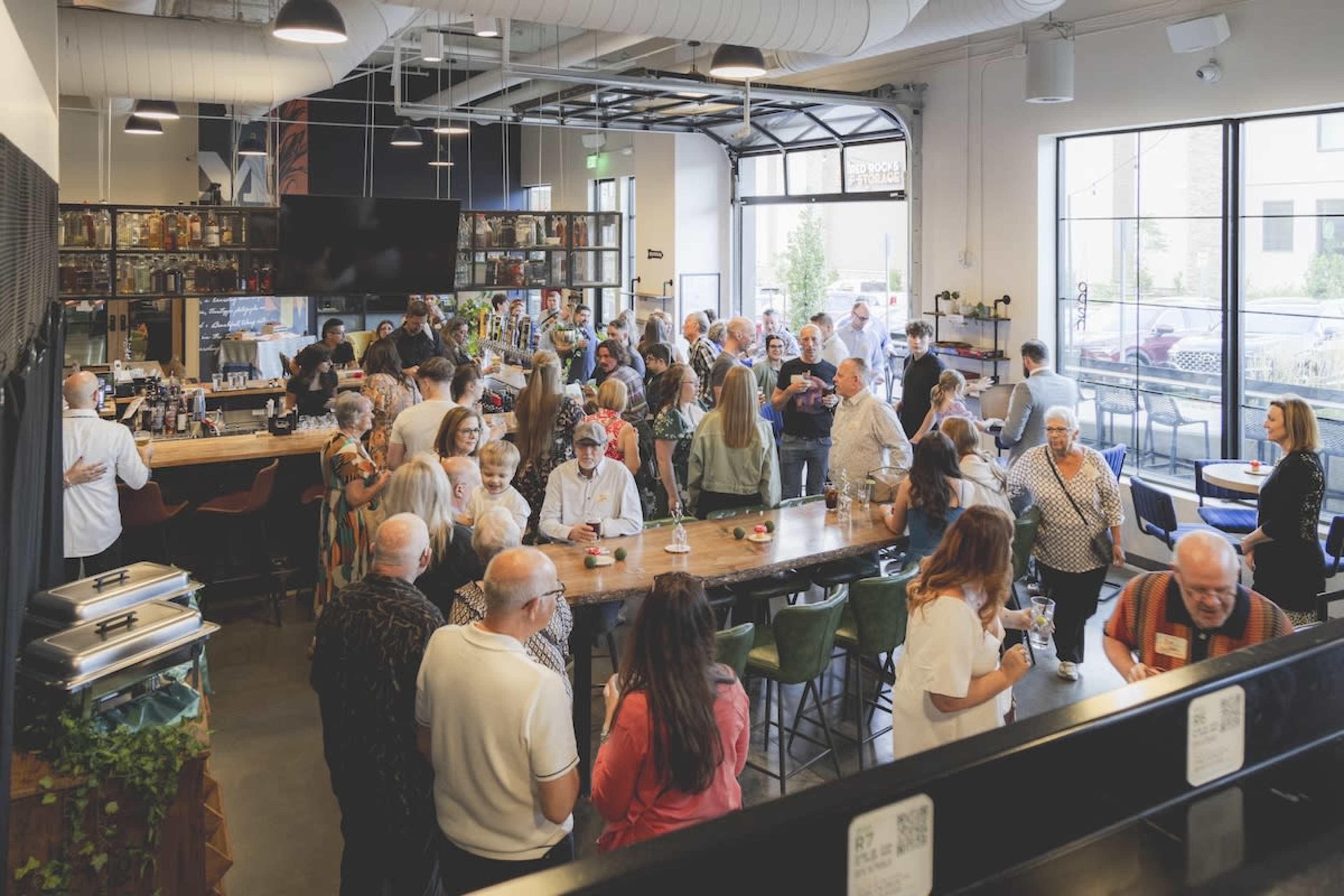 A large group of people gathers in a spacious bar area, engaging in conversation and socializing around a central wooden table.