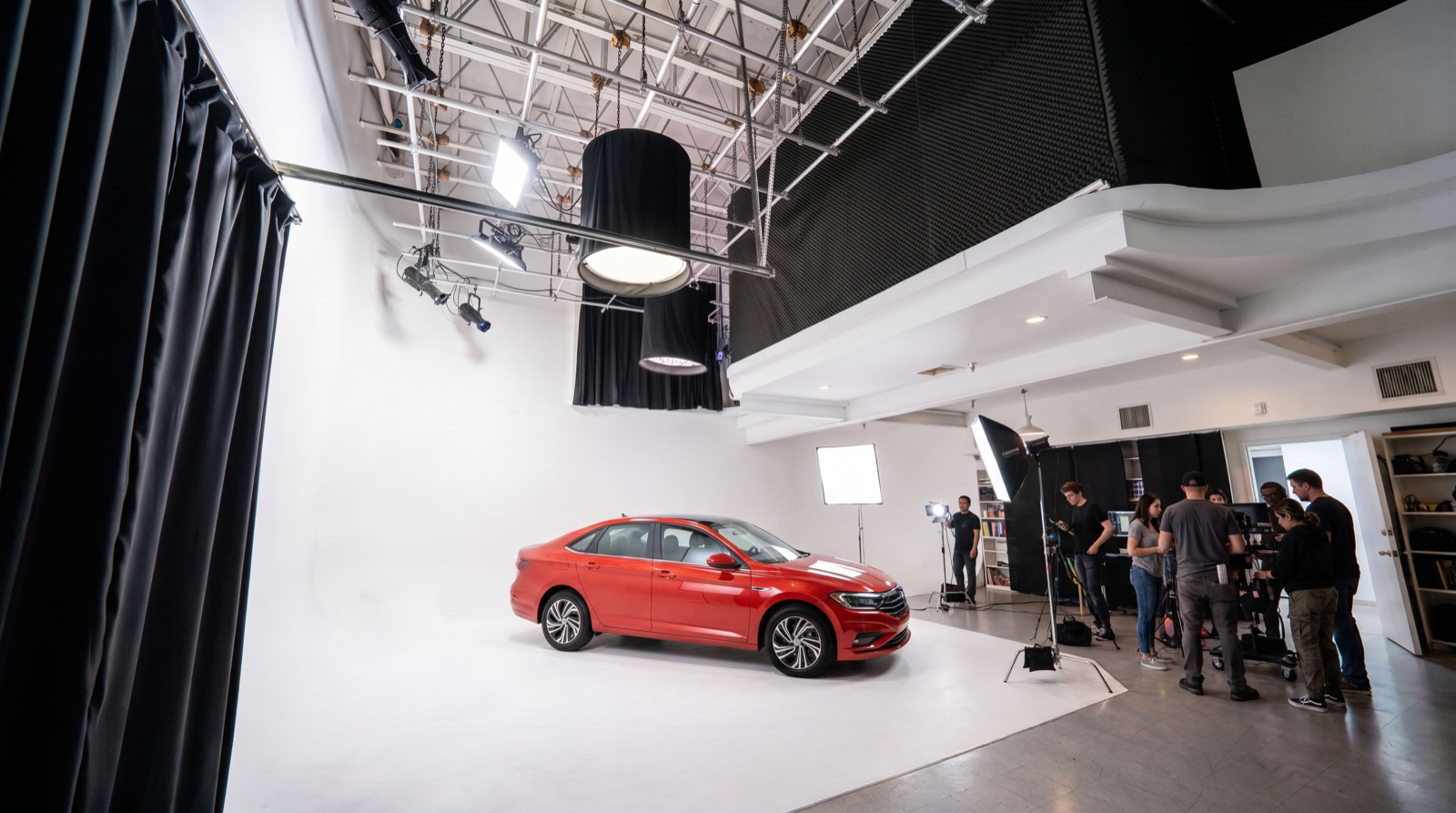 A red car is positioned in a photography studio, surrounded by crew members and studio lights against a white backdrop.