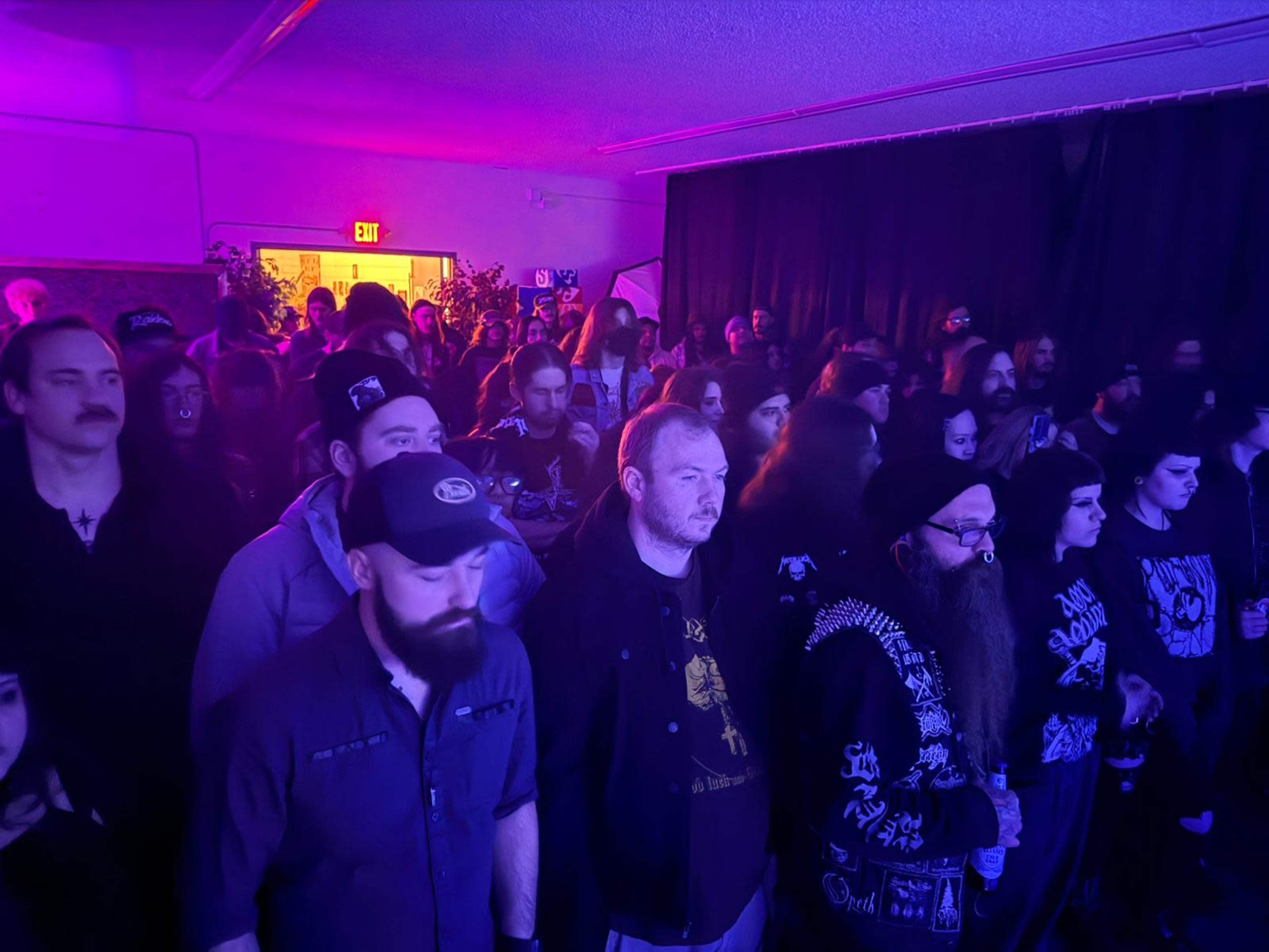 A crowd of people stands close together in a dimly lit space with purple lighting, all facing a stage.