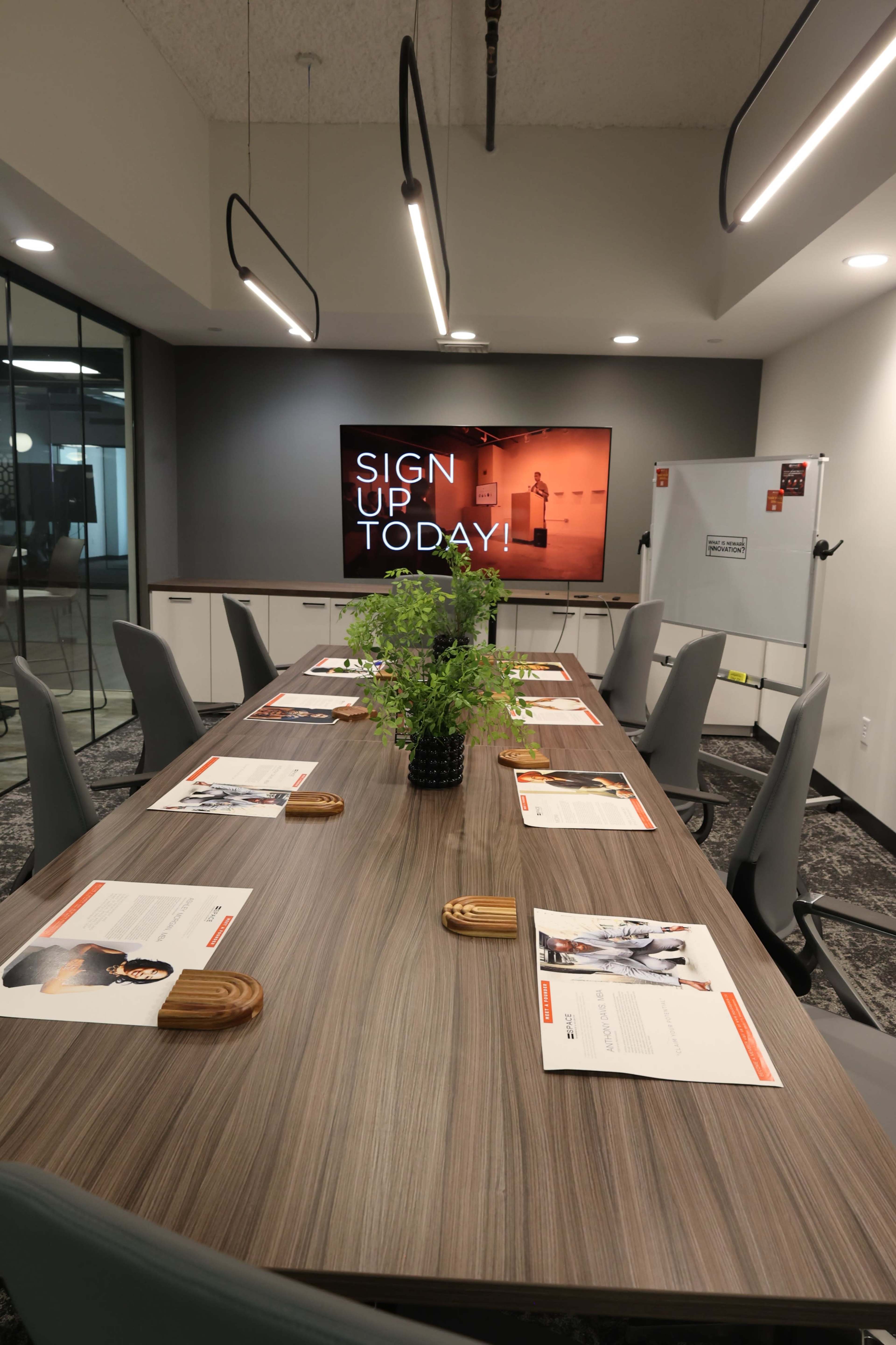 A modern conference room features a large table with chairs, promotional materials laid out, and a screen displaying the message "SIGN UP TODAY!" against a gray wall.