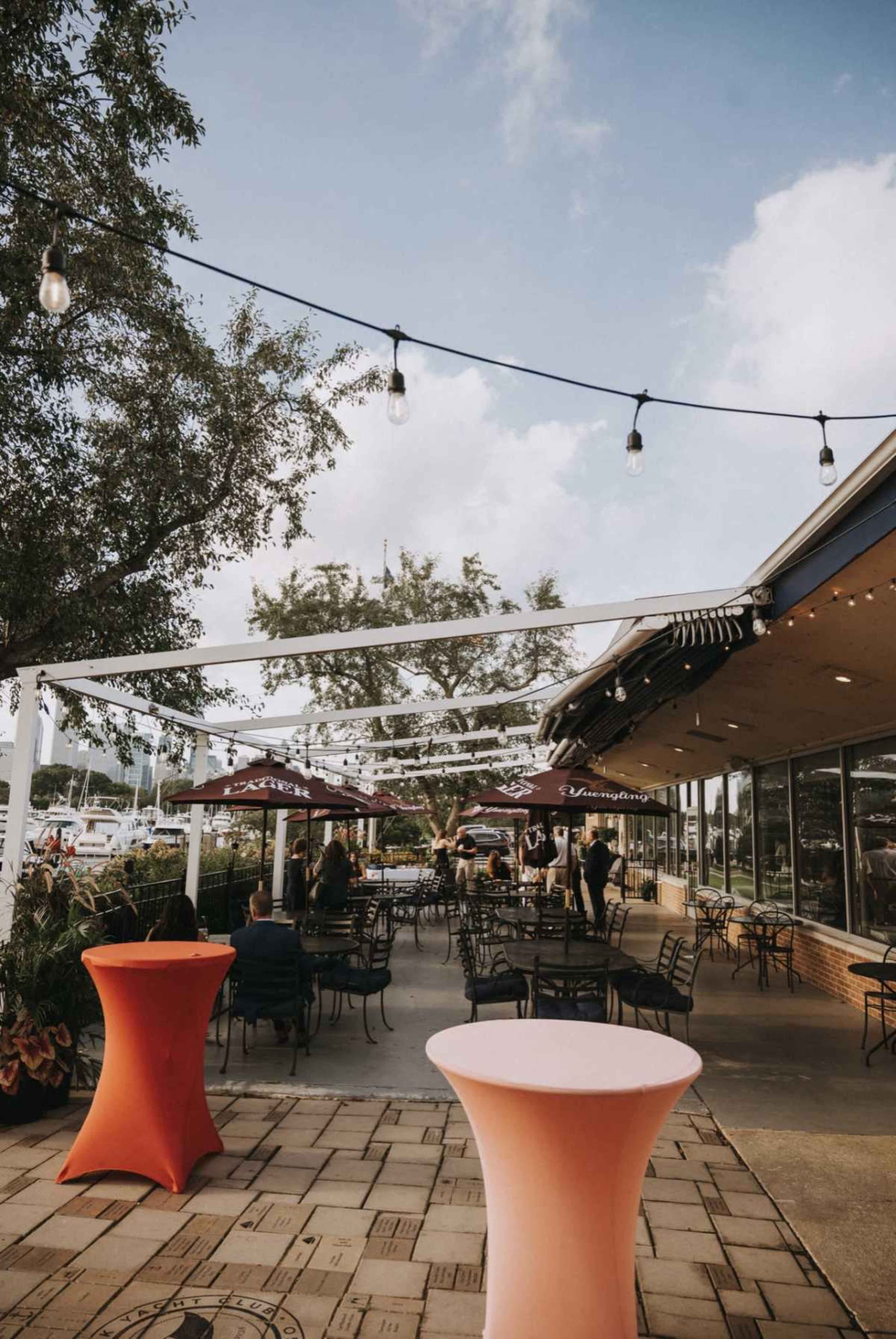 The image shows a patio area of a restaurant with outdoor seating, string lights overhead, and colorful table covers.