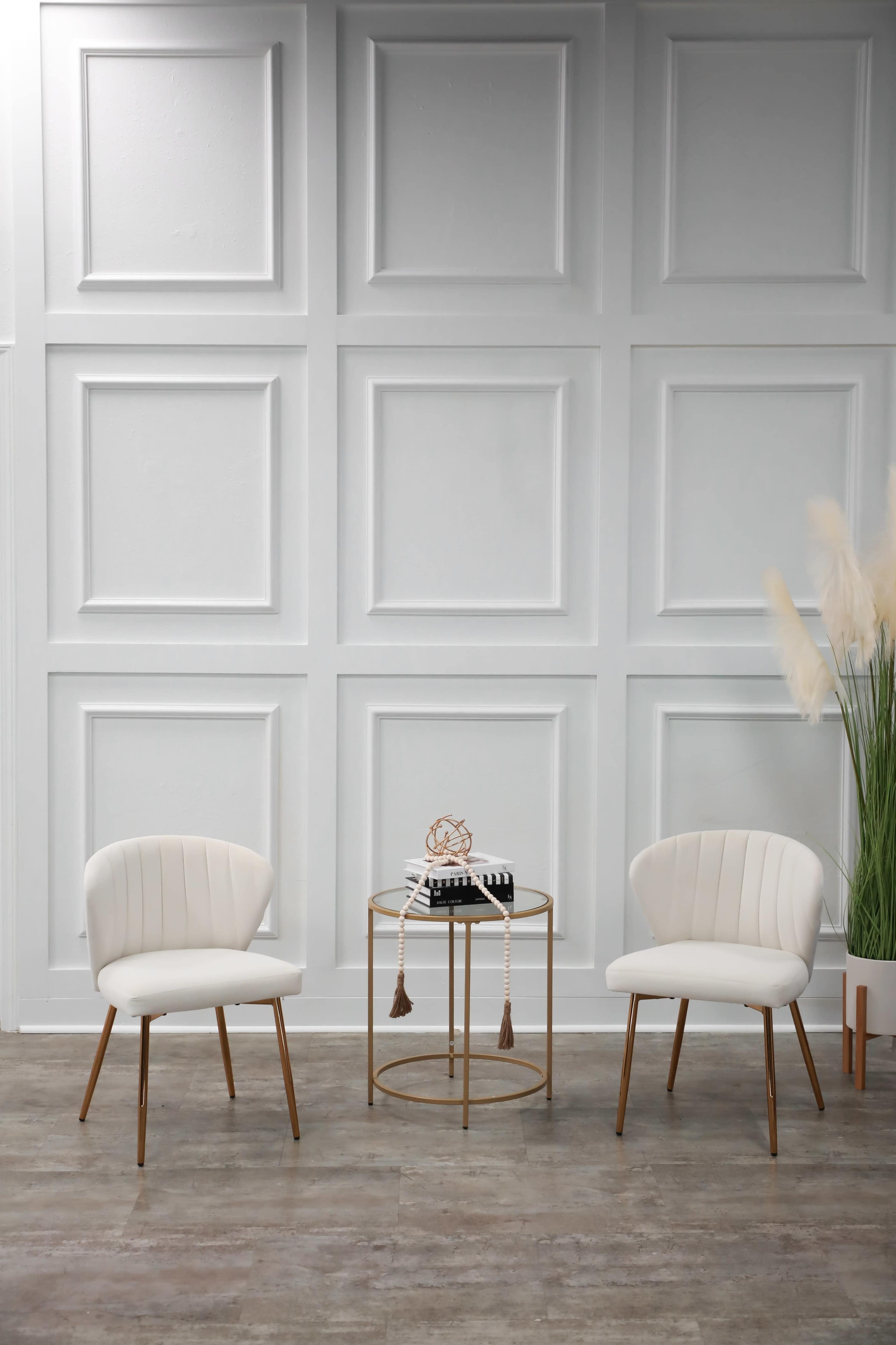 The image features two white chairs and a small round table positioned against a paneled white wall, with tall decorative grasses in a pot beside them.