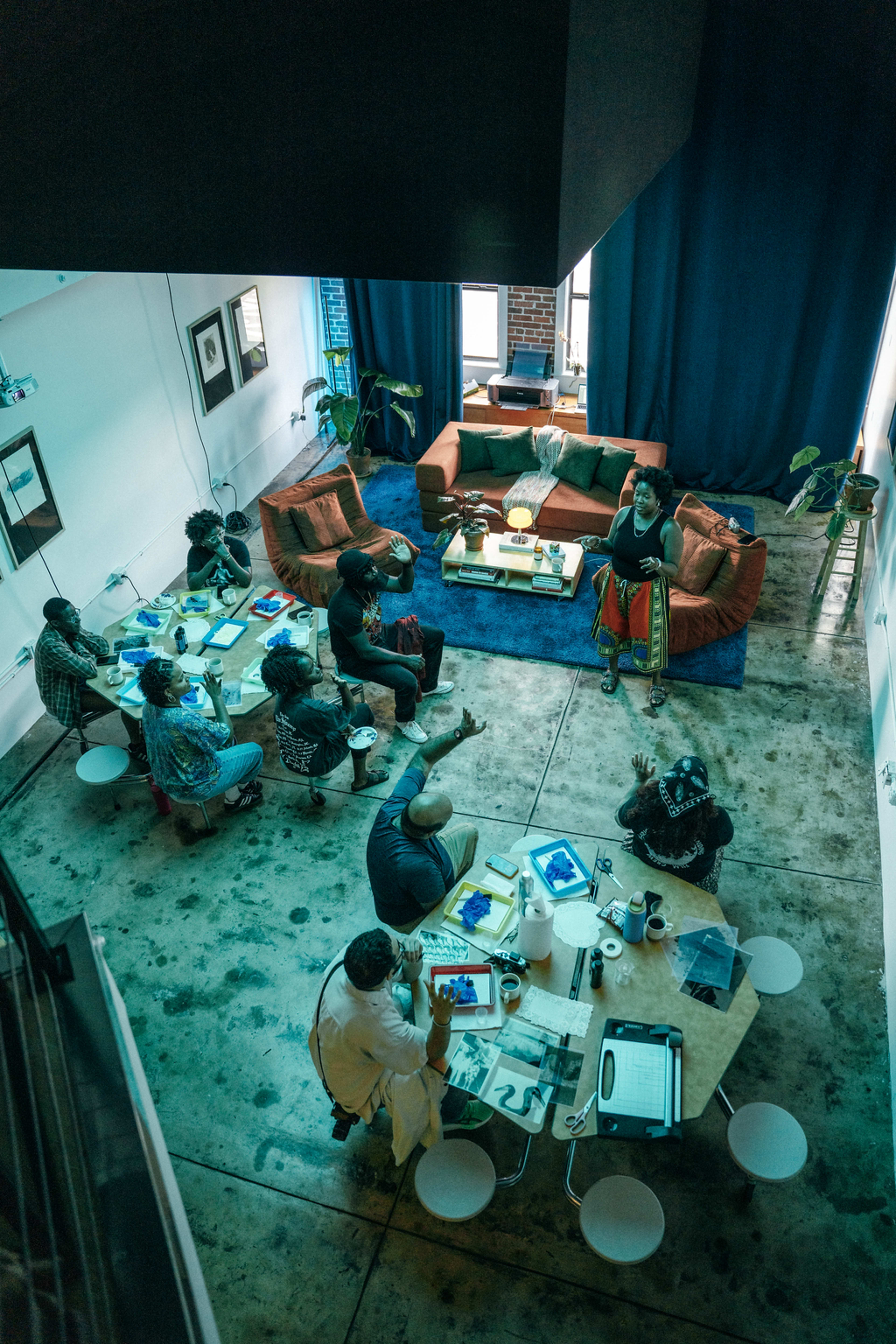 A group of people is engaged in discussion around tables in a well-lit, modern indoor space with a large green couch and blue carpet.
