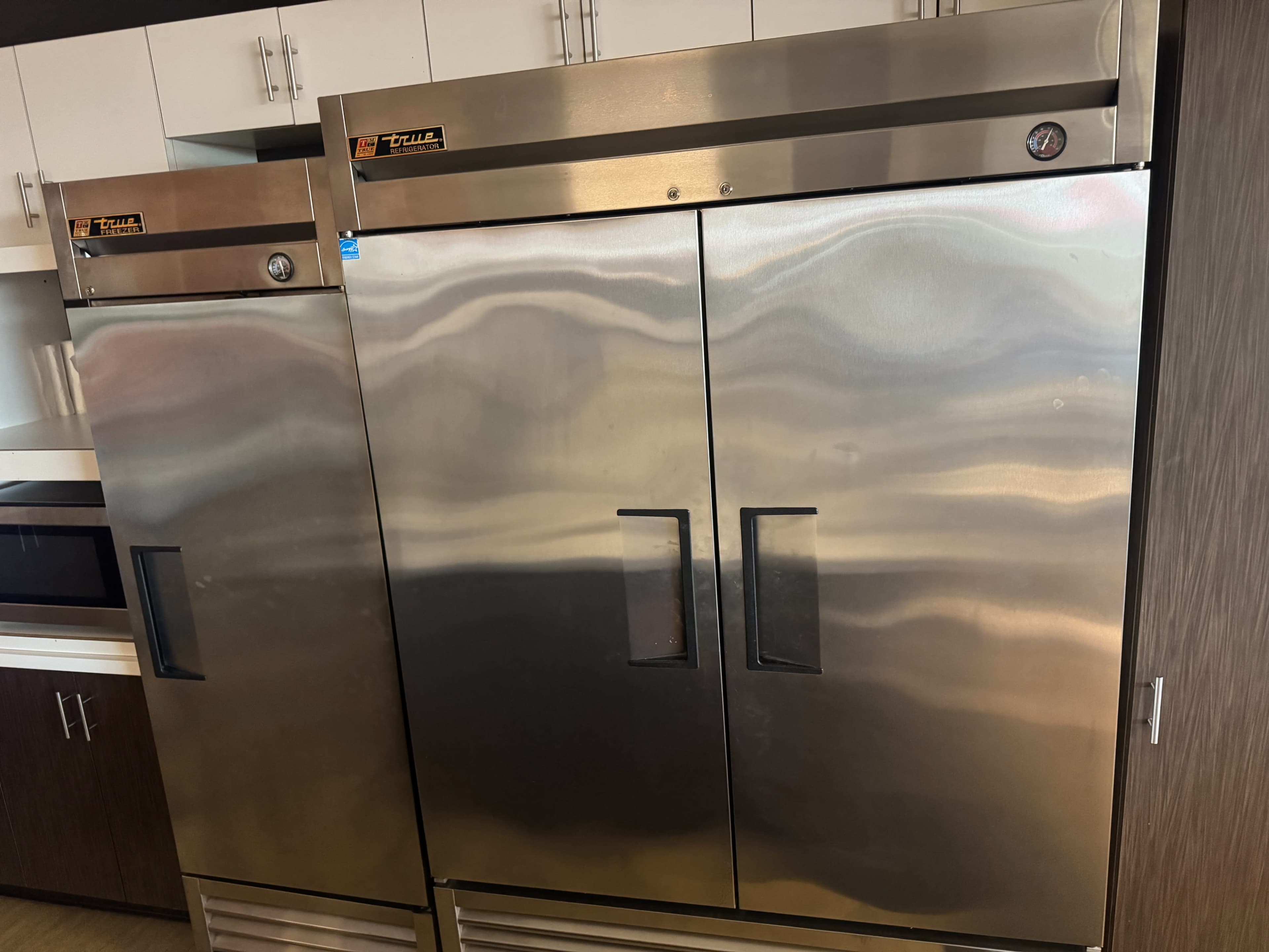 The image shows two stainless steel commercial refrigerators side by side in a kitchen setting.