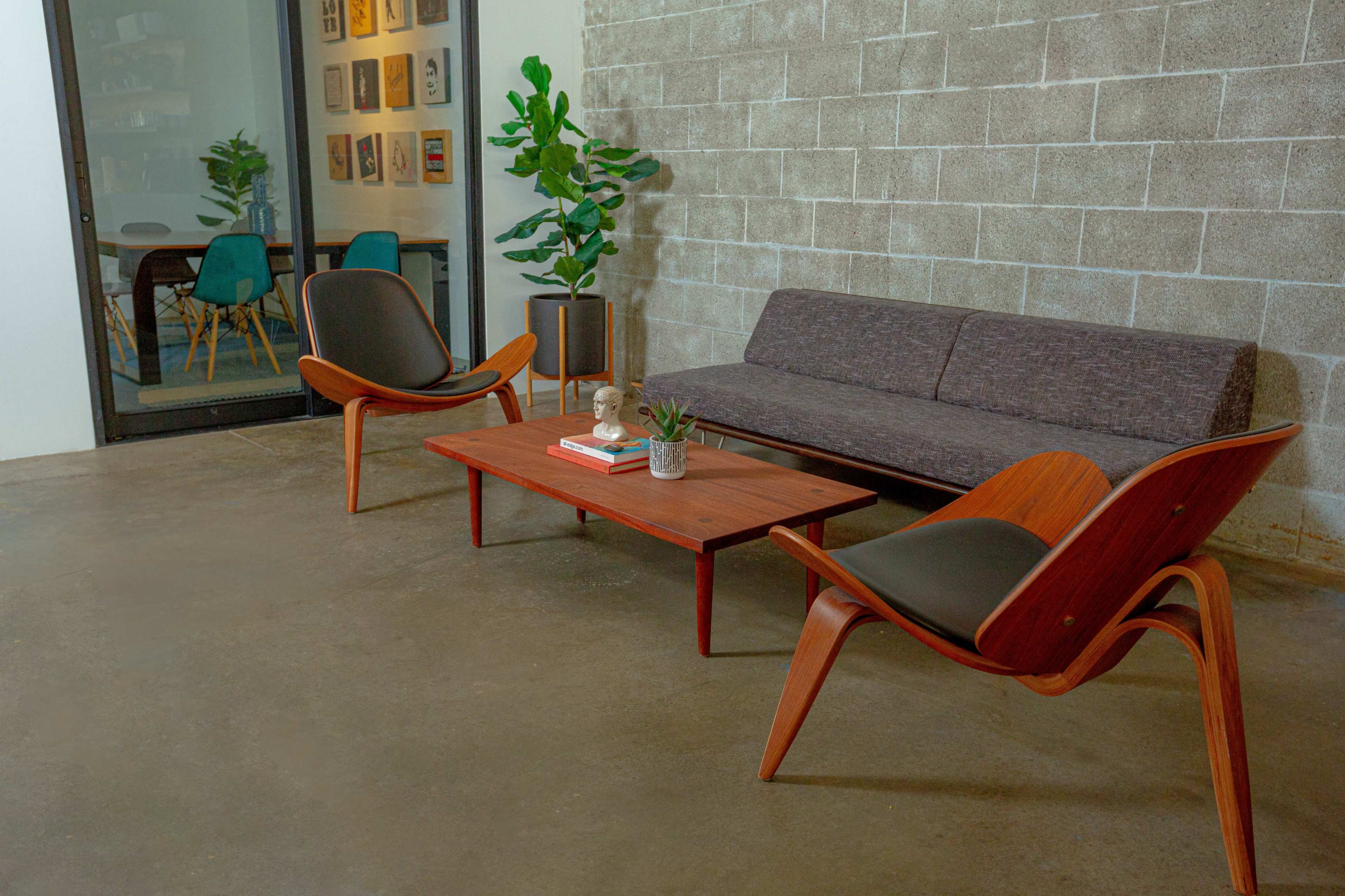 The image shows a modern seating area with a dark sofa, two wooden lounge chairs, and a coffee table, situated against a concrete wall.